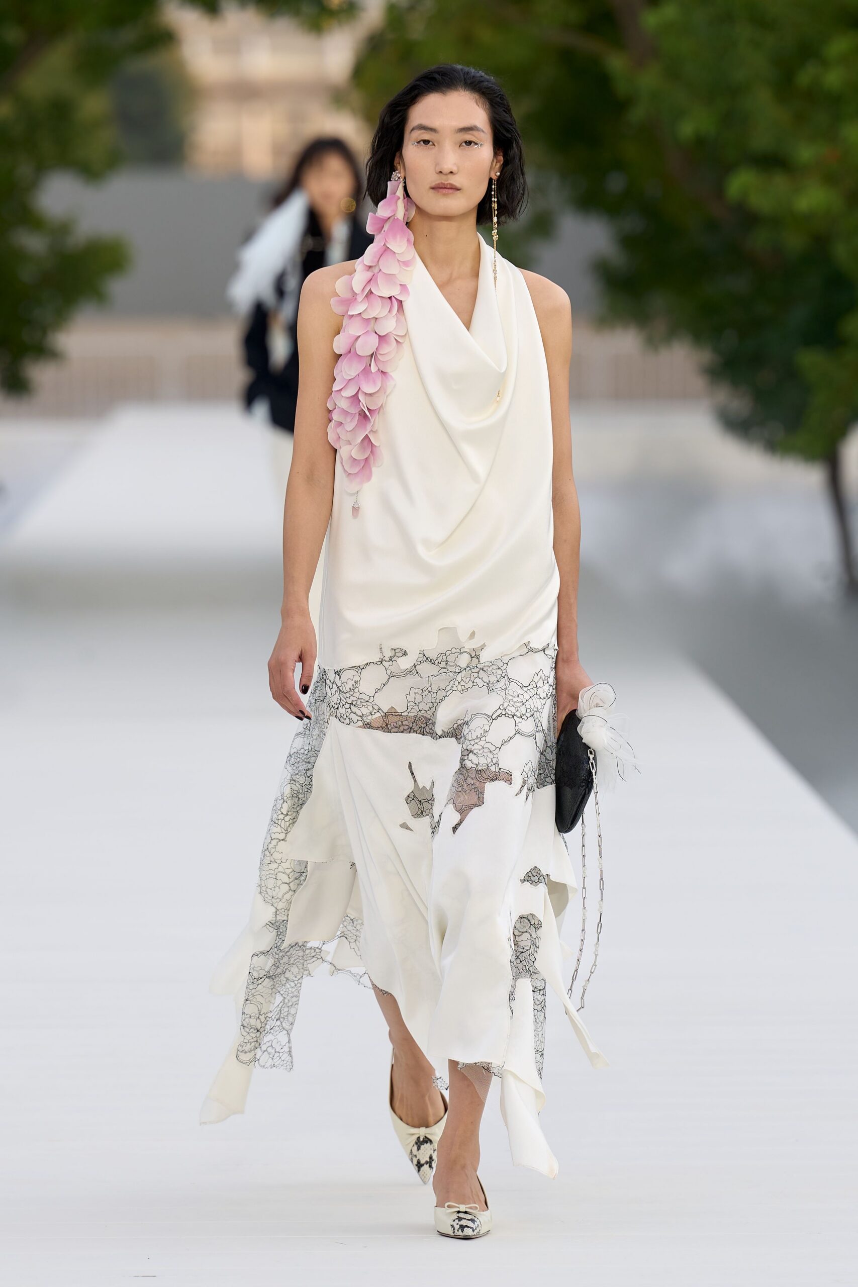Ivory cowl-neck dress in liquid satin, black lace panels breaking the hem. Cascading pink petal garland pinned down one bare shoulder. Black oval clutch with a white organza rosette and chain. Cream pointed pumps with a tiny bow and snake-print toe. Crossing a sculpture garden just after the wedding meal moved outside.