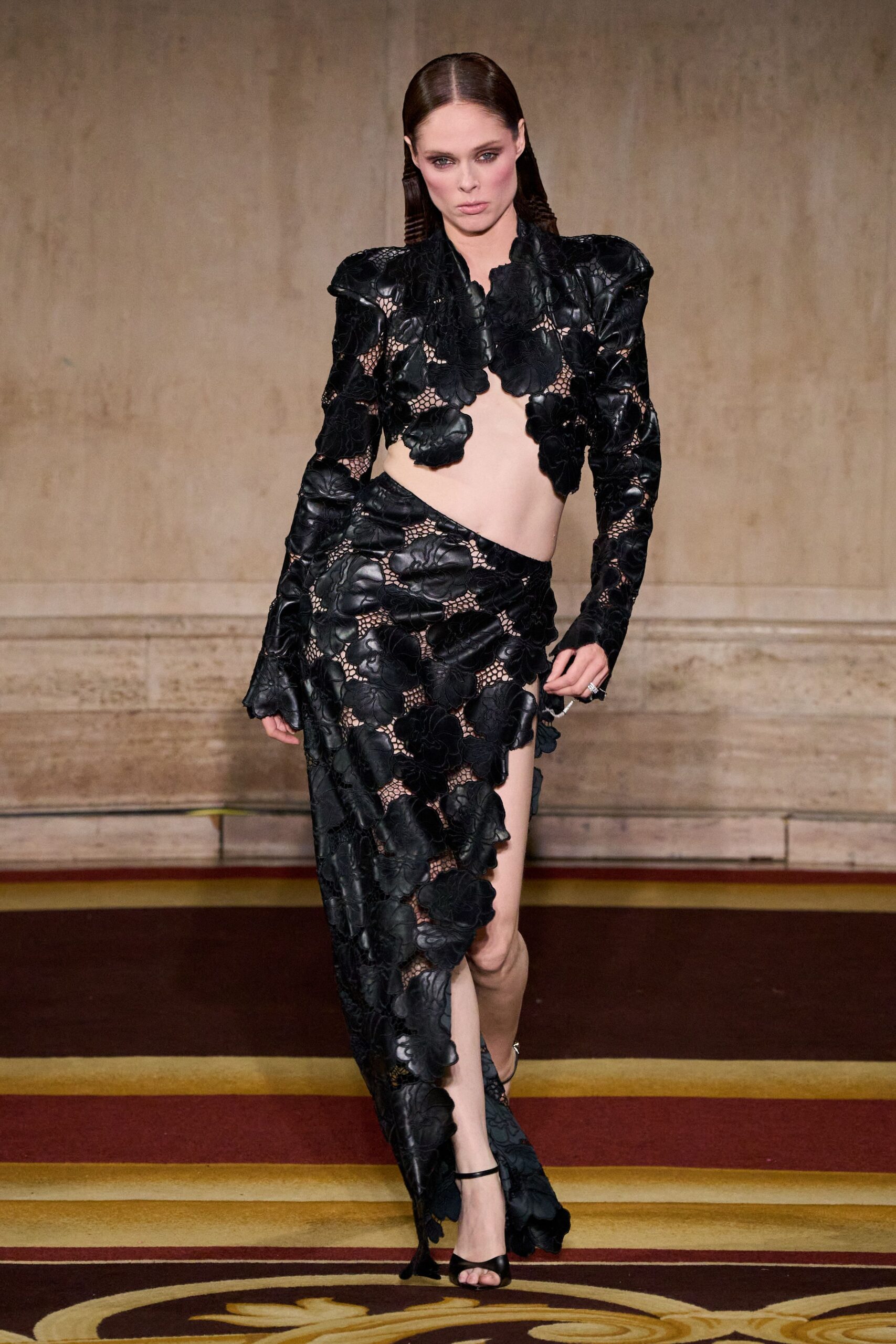 Black leather flower appliques over open net, cut high off the ribs. Matching leather-petal skirt wrapped diagonally, slit climbing to the upper thigh. Black ankle-strap sandals with a narrow peep toe. Crossing a hotel ballroom carpet before anyone has cleared the glasses.