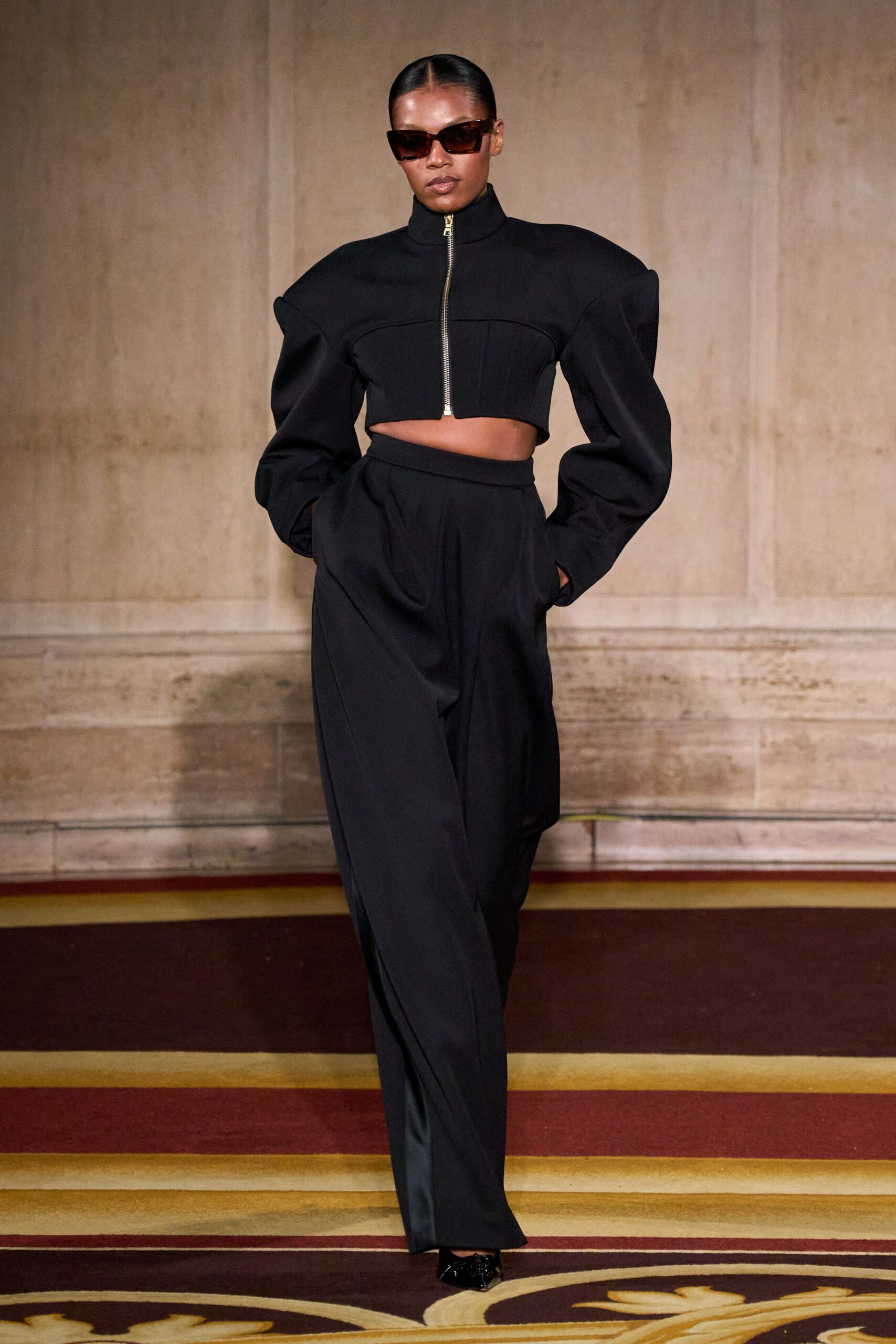 Oversized brown square frames with a slight upward tilt. Black zip-front jacket, funnel neck and arched seam over the bust. High-waisted black trousers with deep pleats and hands sunk in pockets. Black patent pointed pumps. Crossing a hotel ballroom carpet before the awards lunch starts.