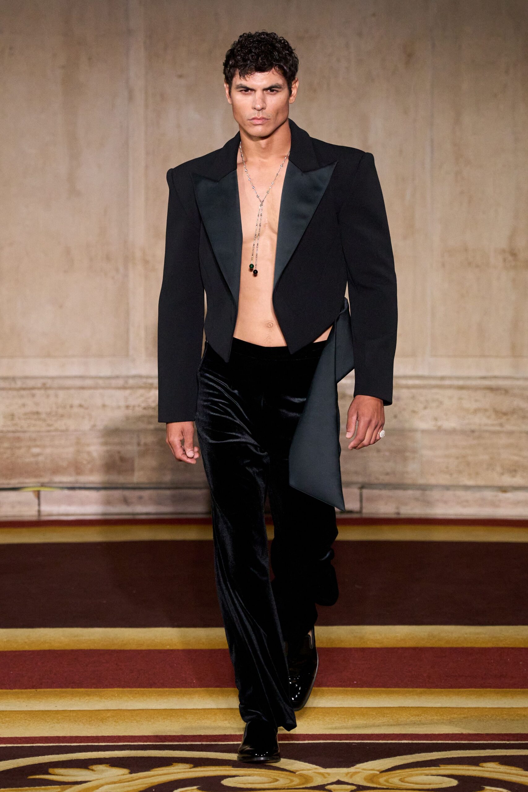 Black cropped jacket with satin shawl lapels and a long side panel. Silver lariat chain ending in two dark beads. Black velvet trousers with a liquid nap catching at the thigh. Glossy black square-toe boots. Crossing a hotel ballroom before the fundraiser doors open.
