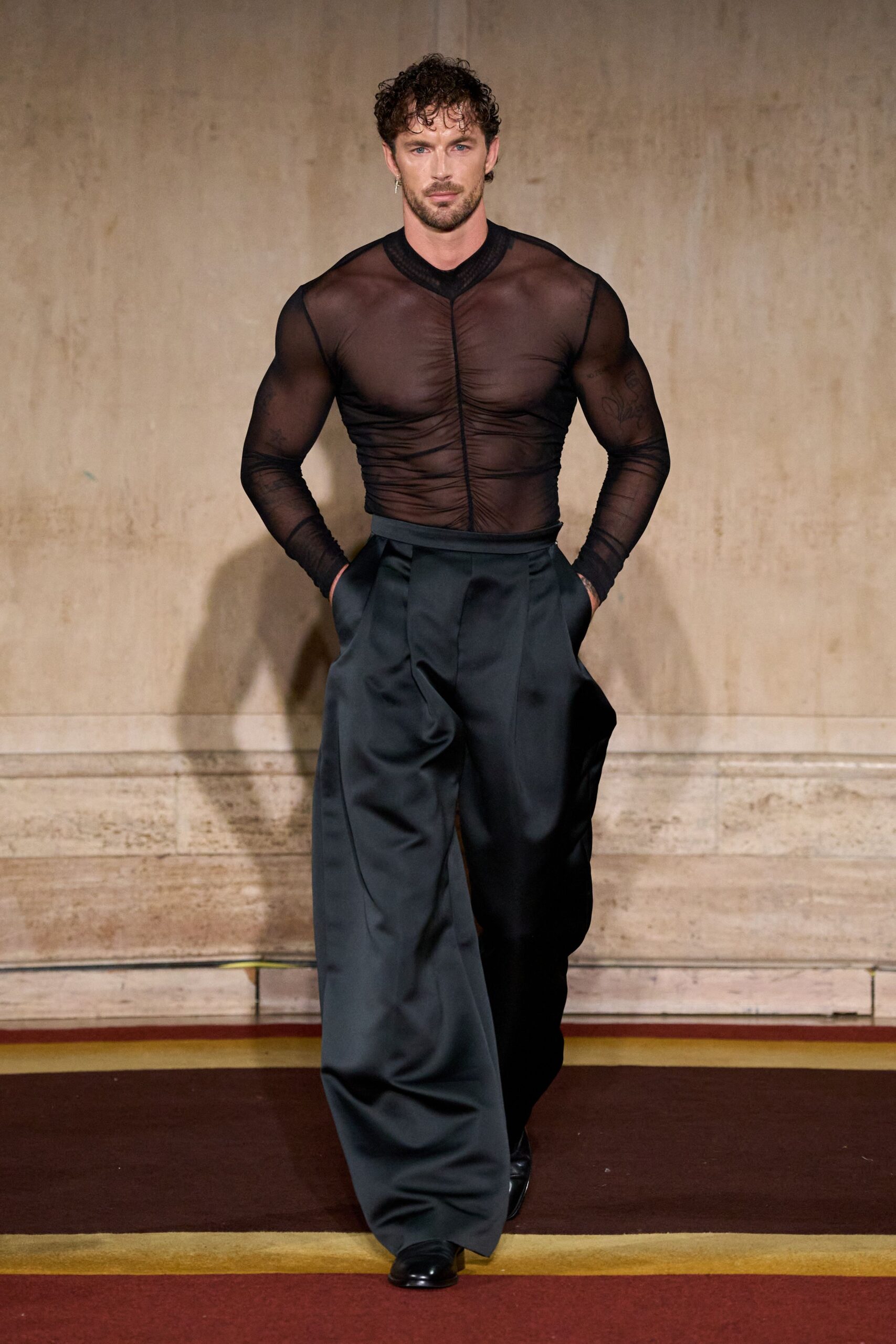 Sheer black mesh top, ruched tight into a vertical center seam. High-waisted charcoal trousers cut extra wide, satin-faced folds breaking over the shoe. Black square-toe boots. Single slim cross earring. After midnight in a courthouse hallway, waiting for the elevator alone.