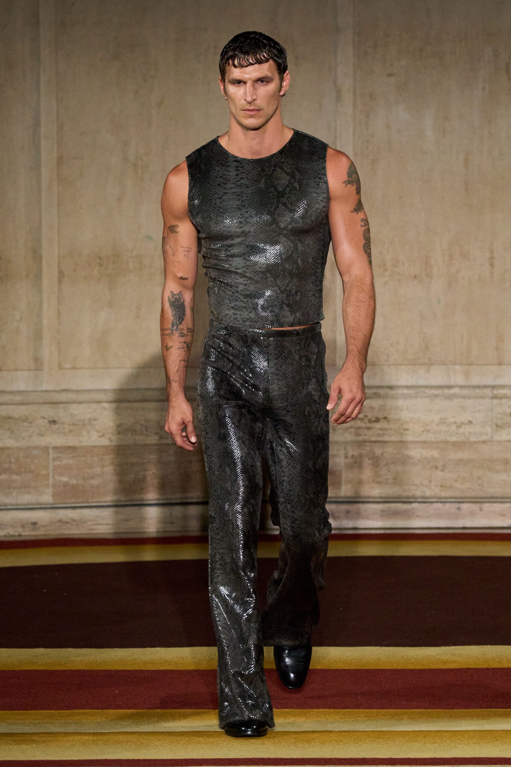 Sleeveless charcoal python-textured leather, cut close with a raw-looking armhole. Matching python-textured leather trousers, straight through the leg with a glossy catchlight. Black square-toe boots peeking under the hem. Hotel corridor carpet at 2pm, still carrying last night's heat.