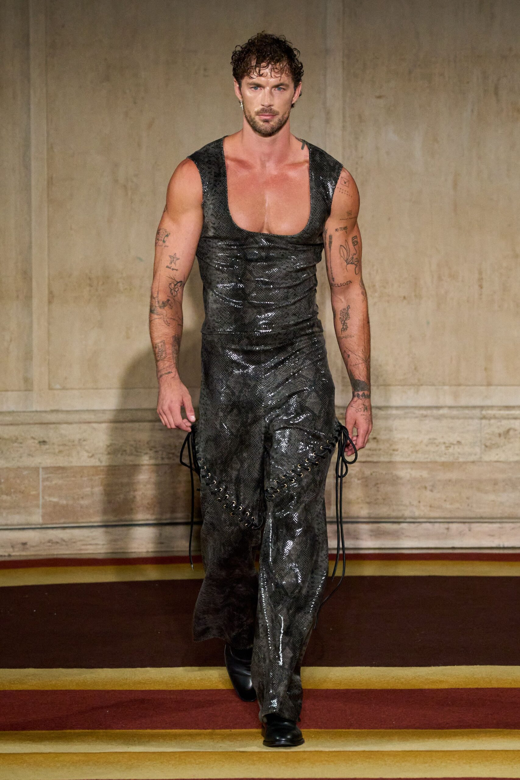 Deep scoop tank in glossy charcoal python-patterned fabric, clinging through the torso. Matching flared trousers with brass-ring lacing curving across both thighs, cords hanging loose. Black leather boots with a blunt square toe. Walking out of a basement club into heat already rising off the pavement.