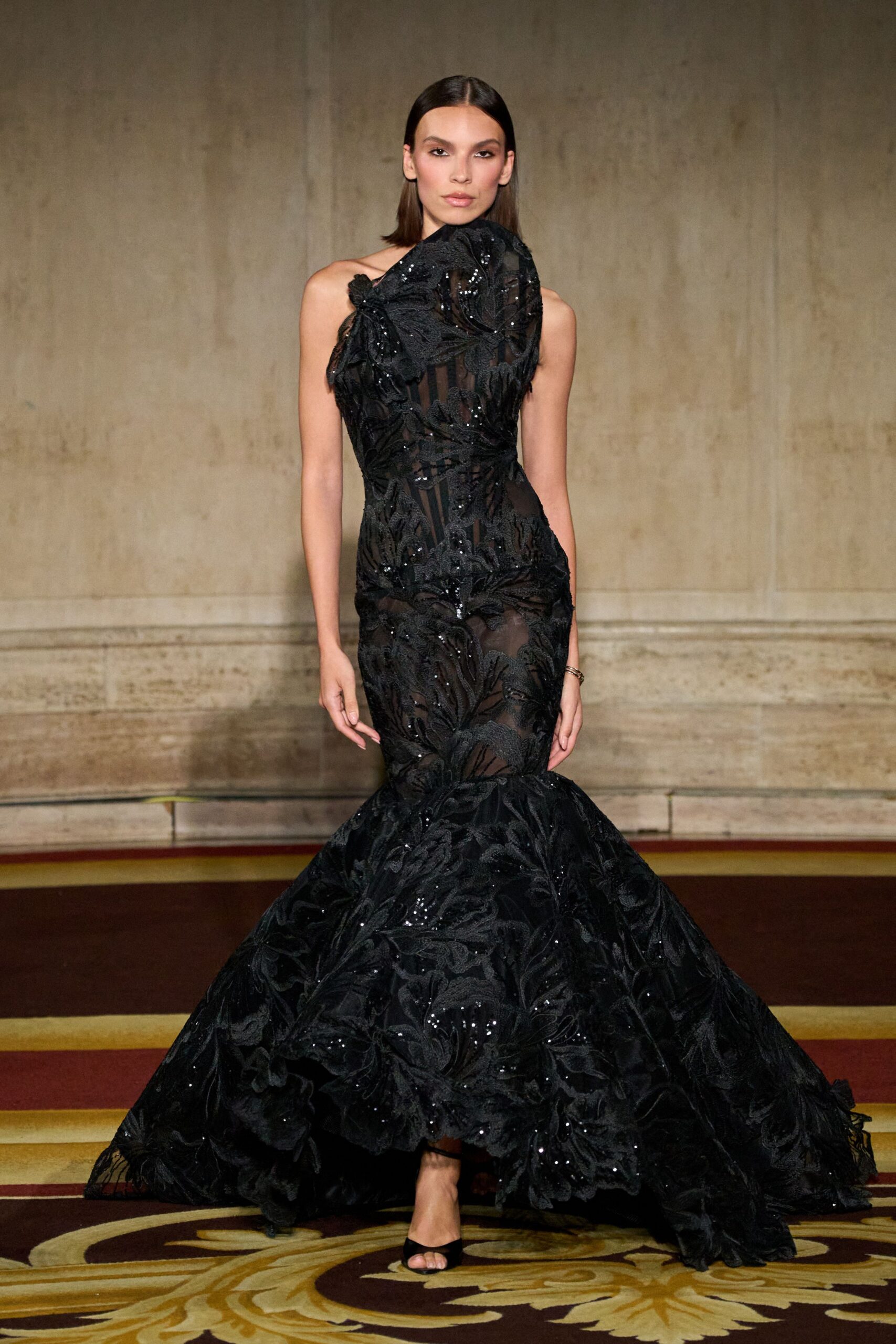 Black one-shoulder mermaid gown, dense leaf embroidery over sheer striped tulle. High sculpted neck panel blooming from the shoulder in matching applique. Black peep-toe mules, barely visible under the flared hem. Slim silver cuff at one wrist. Crossing a hotel ballroom after the orchestra has packed up.
