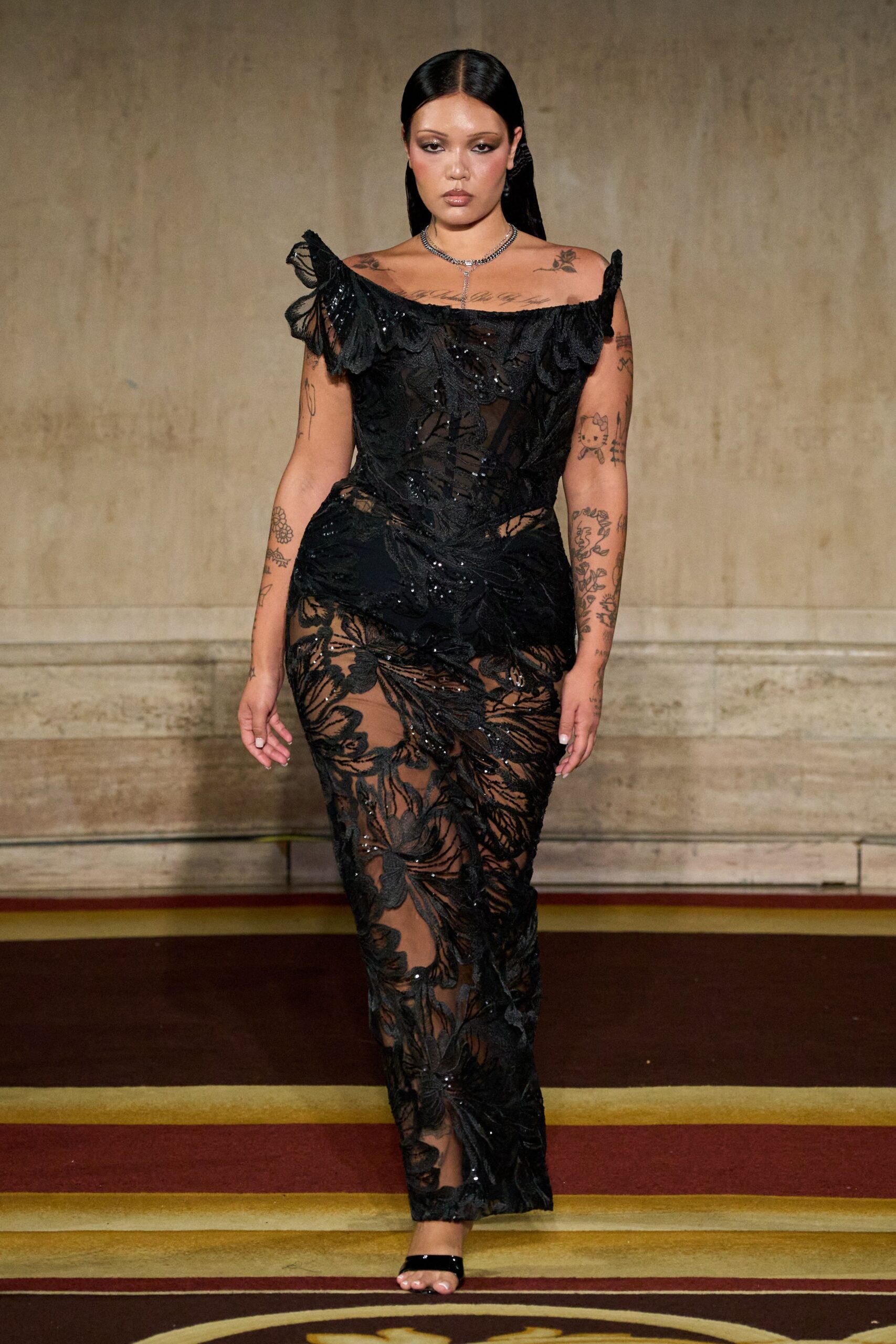 Black illusion gown with dense leaf embroidery and sequins, neckline slipping off both shoulders. Silver Y-necklace with a narrow drop at the sternum. Black patent thong sandals, barely there under the hem. Crossing a faded hotel ballroom carpet after midnight rain.