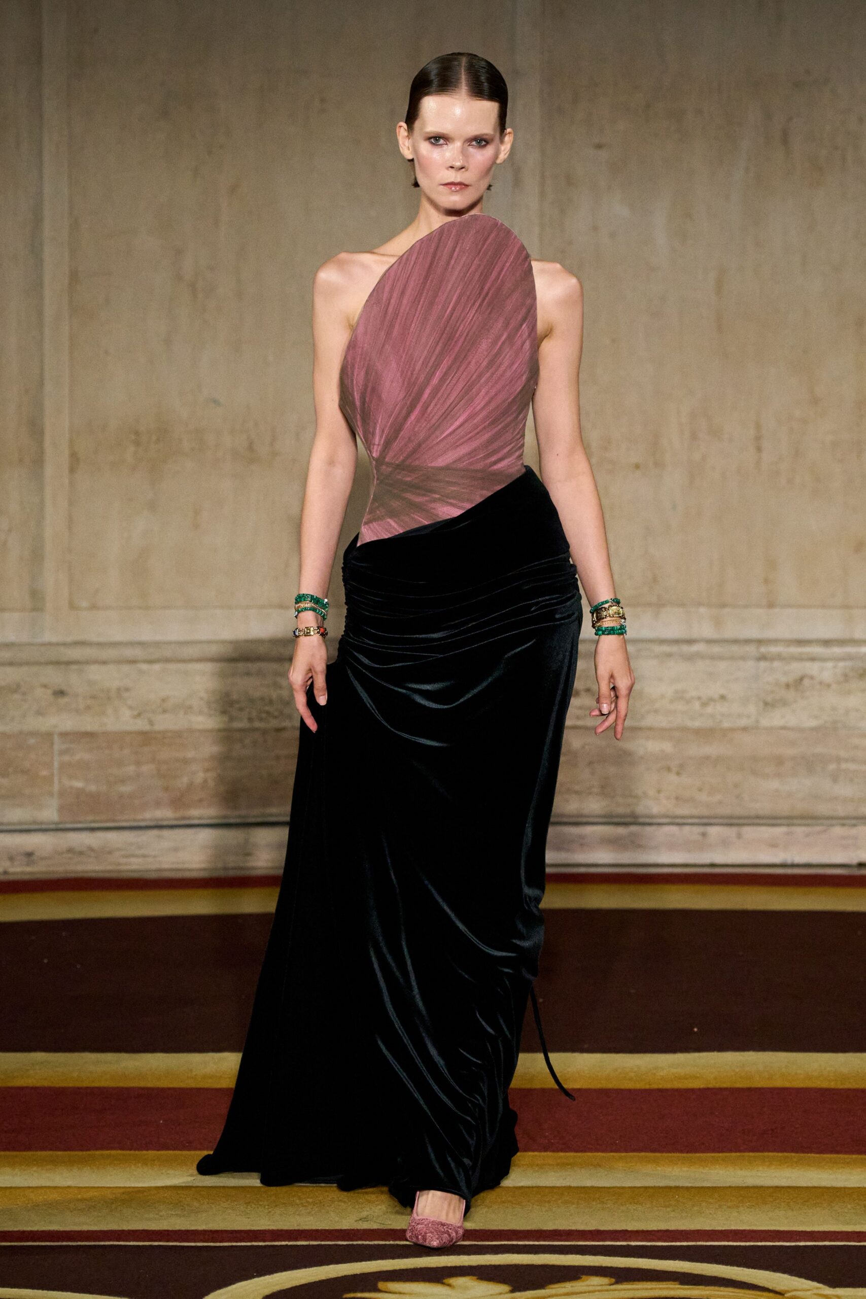 Dusty rose ruched bodice rising into a tall asymmetric petal panel. Black velvet floor-length skirt, densely draped across the hips. Stacks of green bangles and one chunky gold cuff. Rose textured pumps peeking from the hem. Crossing a hotel ballroom carpet before the string quartet starts.