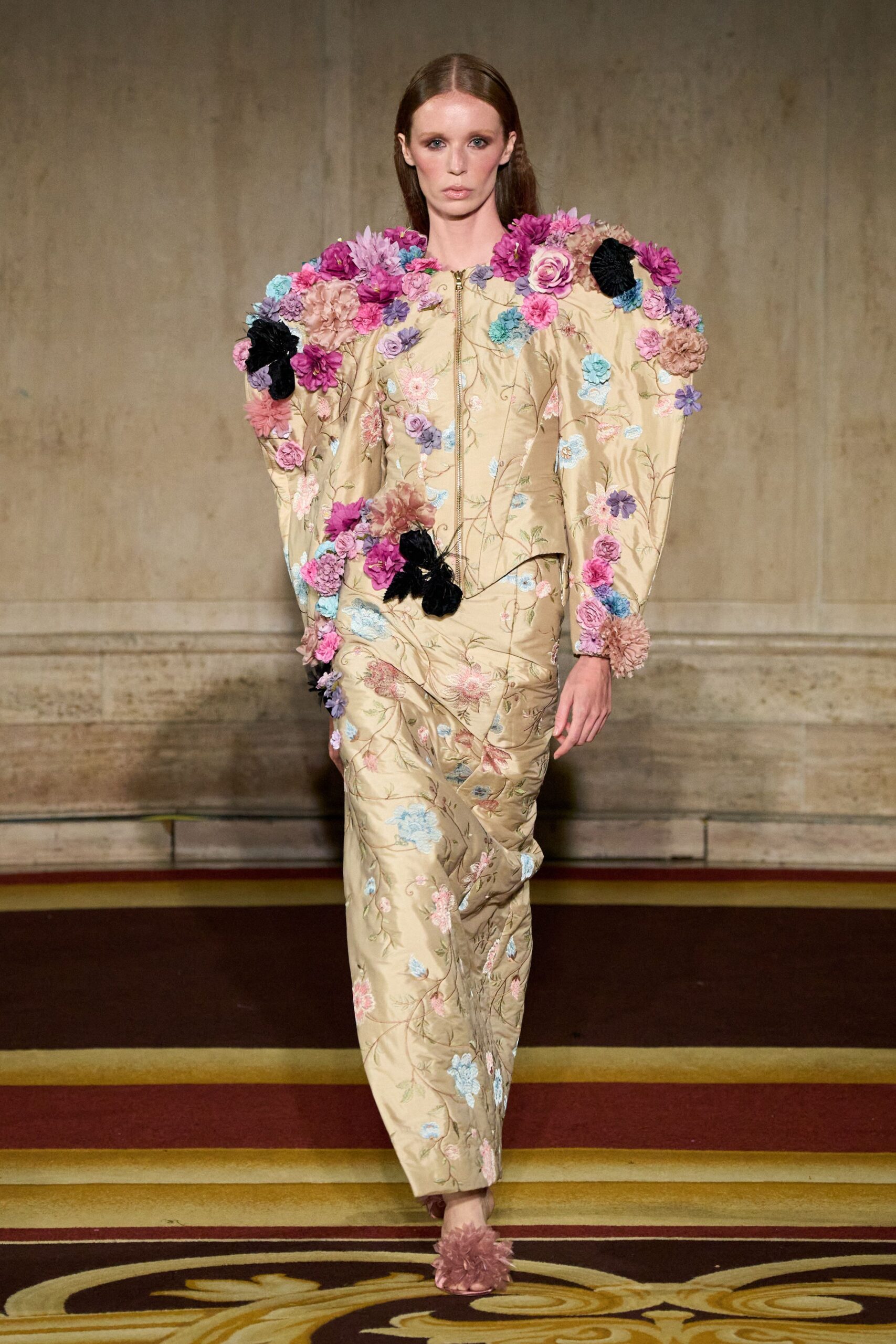 Champagne brocade jacket with ballooned sleeves, crowded in wired rosettes. Matching column skirt in trailing floral brocade, slightly ruched at the hip. Dusty rose mules topped with a crushed tulle pompom. Crossing a hotel ballroom after the wedding cake has been cleared.