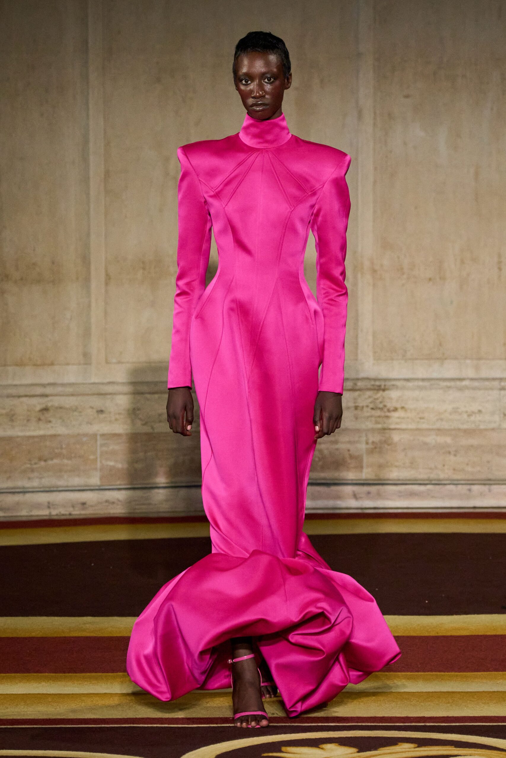 Fuchsia satin gown with a high funnel neck and sharp padded shoulders. Barely-there ankle-strap heels peeking beneath the pooled fishtail hem. Crossing a hotel ballroom carpet after midnight, still carrying the room's silence.