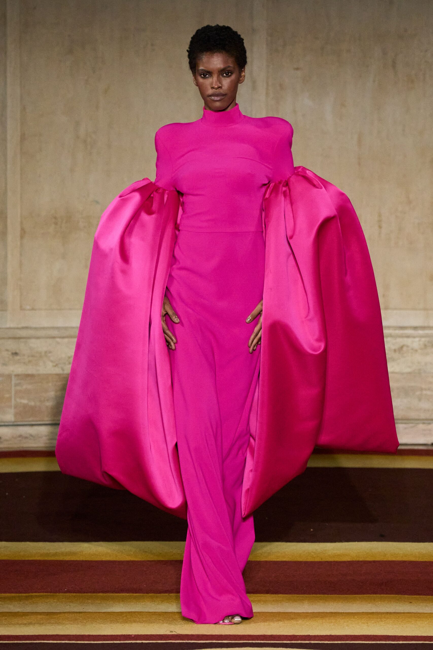 Fuchsia stretch column gown with a high mock neck and sculpted hips. Gigantic satin cape sleeves knotted at the upper arm, ballooning to the floor. Matching pink sandals, only the front sliver peeking out. Descending a hotel staircase after midnight, wrapped in banquet-chair satin.