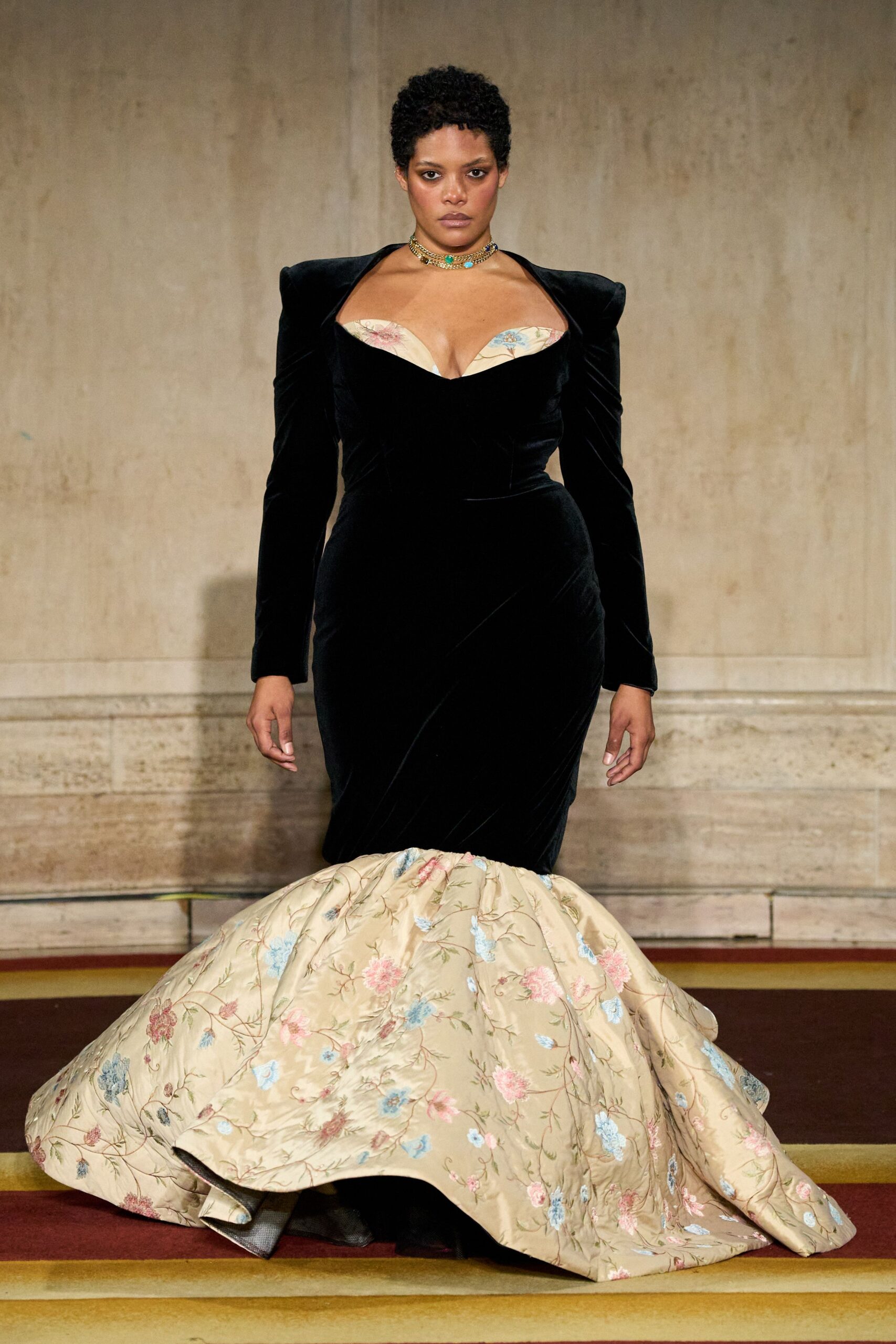 Black velvet mermaid gown with squared shoulders and a floral brocade flounce. Stacked gold chokers dotted with turquoise and cobalt stones. Crossing a municipal ballroom floor after the orchestra has packed up.