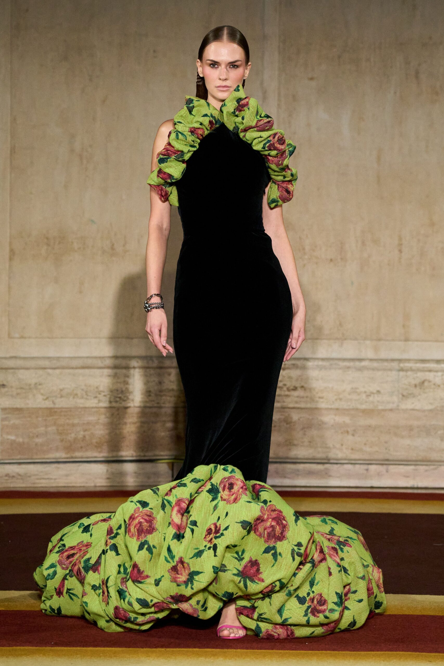 Black velvet column gown with ruched chartreuse rose brocade framing the neckline and train. Stacked gunmetal chain bracelets at one wrist. Hot-pink barely there sandals peeking under the pooled hem. Crossing a ballroom after midnight with the curtains still open to the garden.