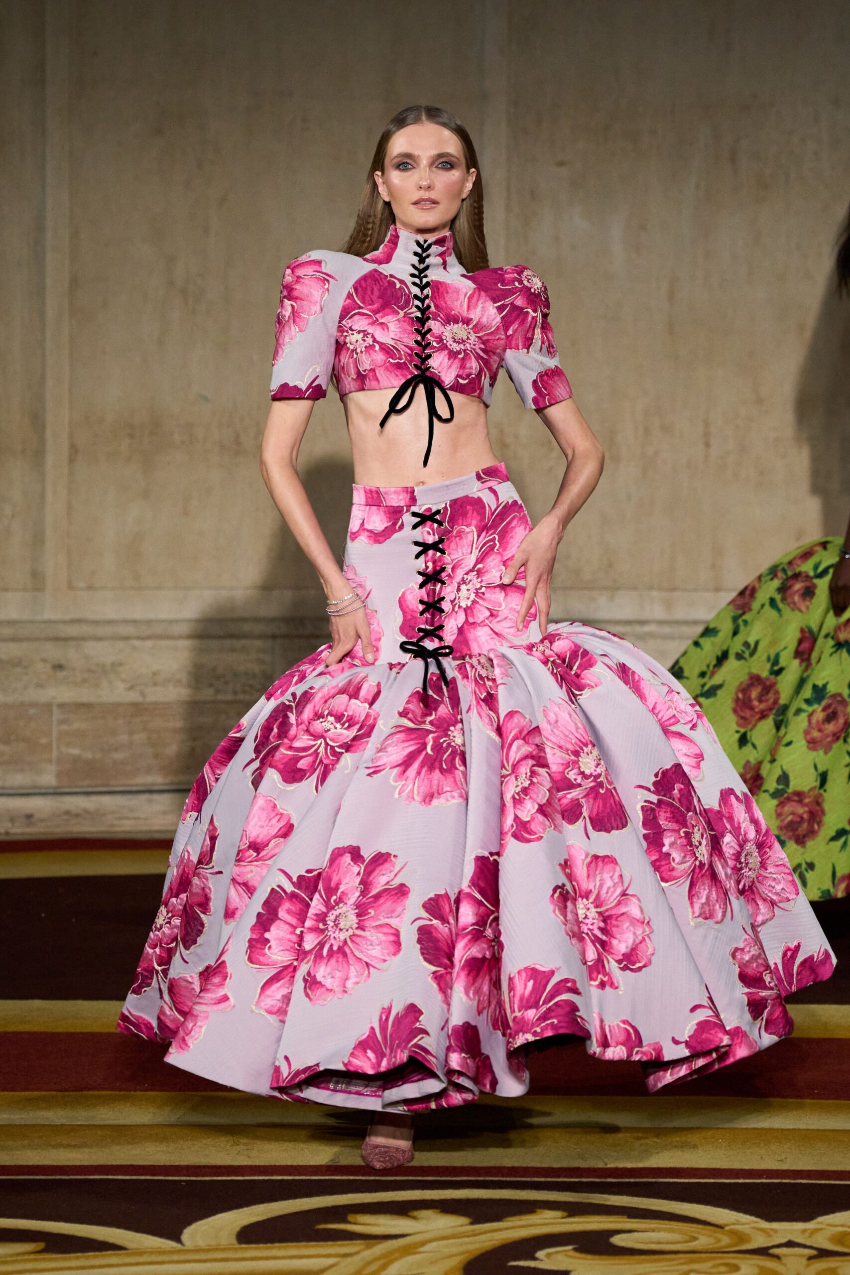 Lilac floral crop top with puff sleeves and black corset lacing. Matching ball skirt, front slit laced low and hem held in deep folds. Pink glitter pumps peeking from under the sweep. Descending a hotel staircase toward a wedding nobody said was formal.