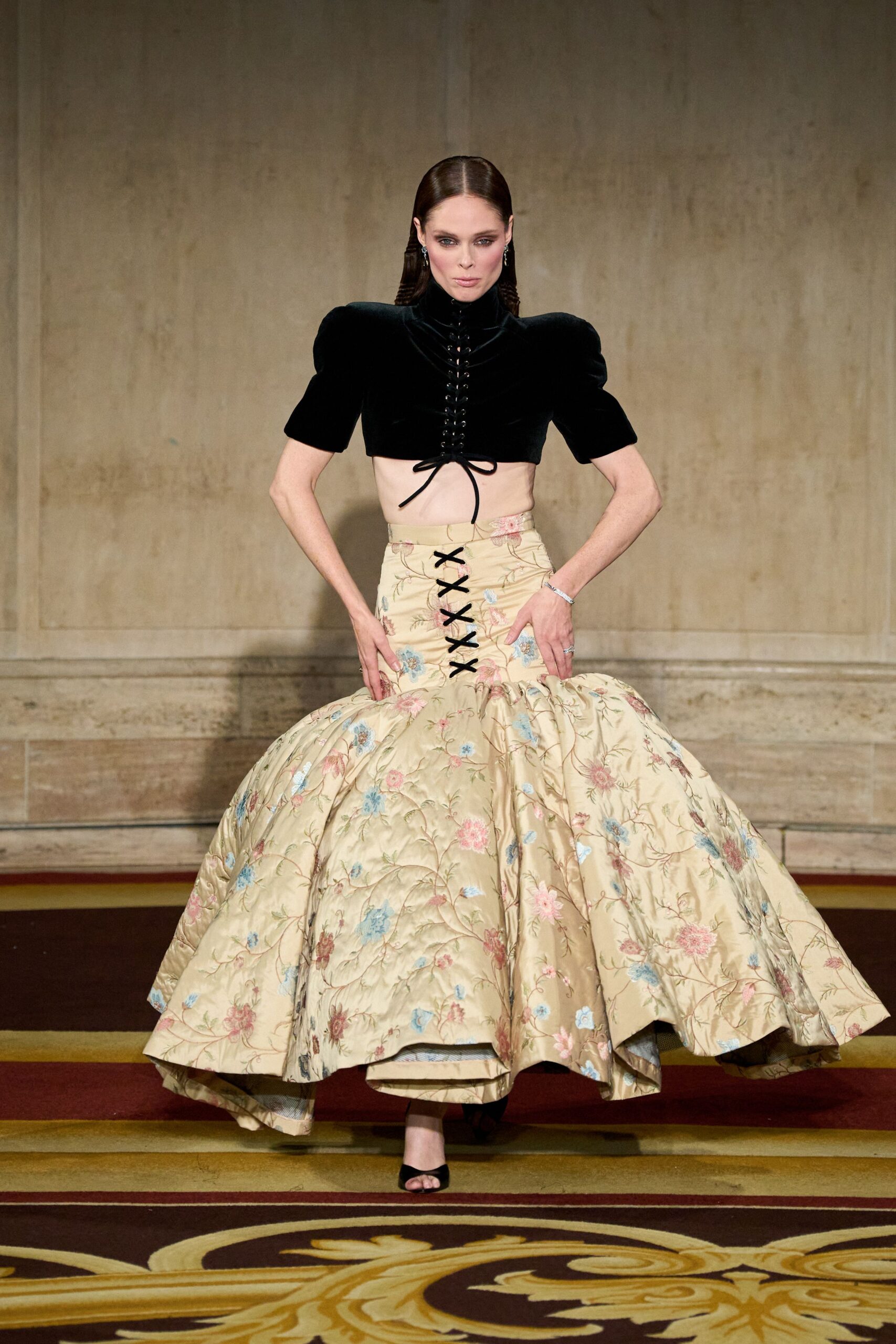 Black velvet crop top with puffed sleeves and center corset lacing. Cream floral quilted skirt, black lacing running down the dropped waist. Black peep-toe mules. Crossing a hotel ballroom carpet after everyone else has gone upstairs.