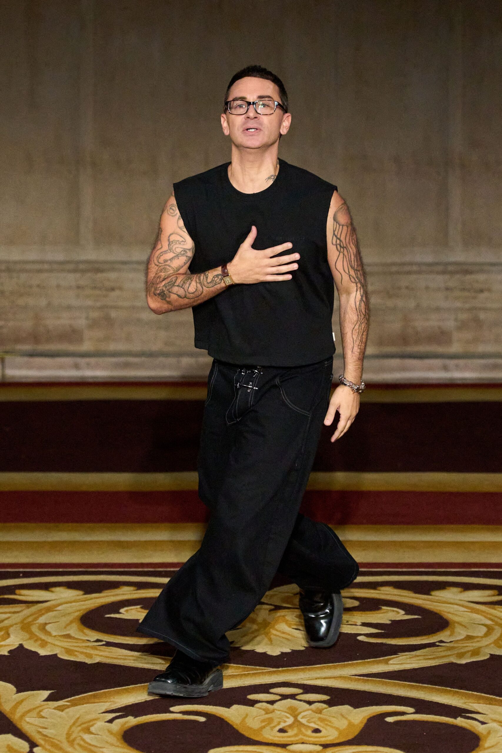 Black rectangular frames with clear lenses. Black boxy sleeveless top, cropped bluntly at the hip. Wide black trousers with contrast topstitching and a laced fly. Square-toe black leather shoes, lightly scuffed at the front. Mixed metal and leather wrist stack. Crossing the hotel ballroom after everyone else has gone to bed.