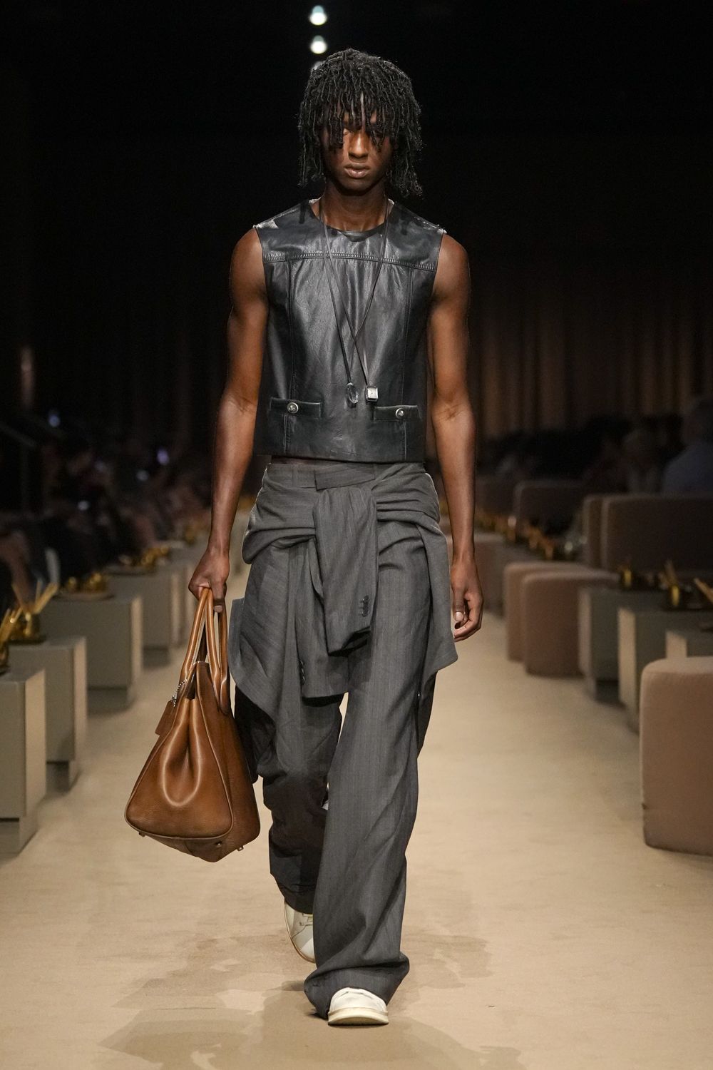 Black leather shell with welt seams and two flap pockets low. Long cord necklaces with a crystal drop and tiny cube. Charcoal wide-leg trousers with a jacket knotted hard at the hips. Cognac holdall, soft sides collapsing into deep folds. White slip-on sneakers. Walking into a hotel lobby after missing the last train.