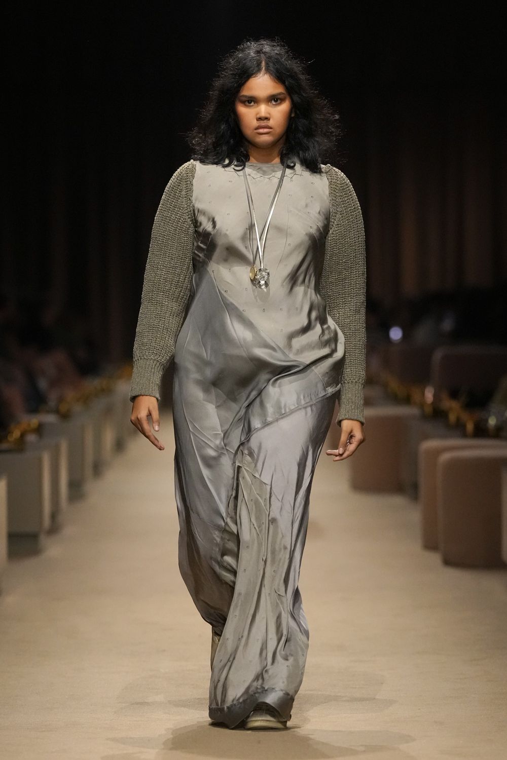 Pewter satin slipdress, bias-draped into a low twisted apron fold. Moss-grey chunky knit sleeves grafted onto the dress bodice. Long cord pendant with one cloudy crystal and one small gilt disc. Dusty taupe toes barely peeking under the pooled hem. Walking across a hotel corridor before the conference rooms open.