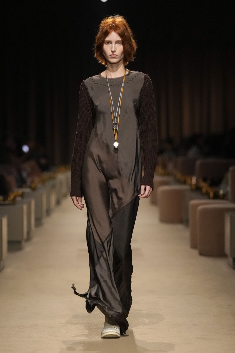 Smoke-brown satin slip dress, sheer hem twisting into a little trailing corner. Espresso open-knit sleeves grafted to the dress, zigzag topstitch at the neck. Long cord necklace with two mismatched pendants hitting the sternum. Pale rubber clogs, blunt and slightly creased. Walking the hotel corridor at dawn for ice and aspirin.