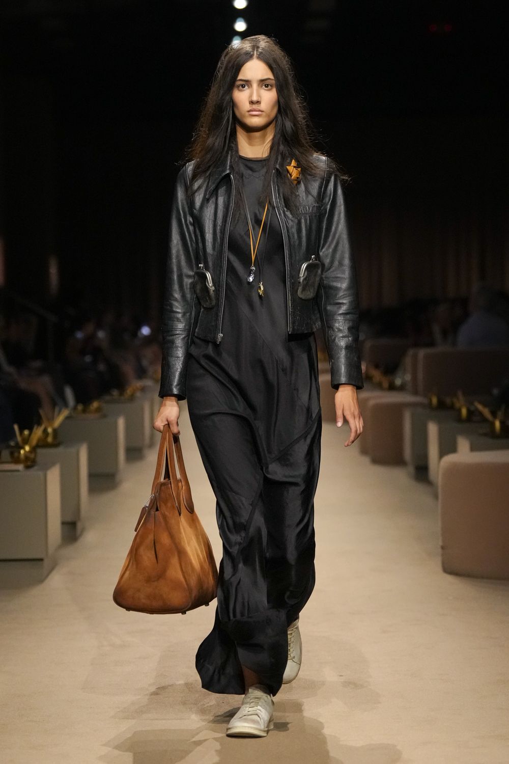 Black leather jacket with clipped pouch pockets hanging at the ribs. Ink satin slipdress cut on the bias, hem pooling over the shoes. Long tan cords with mismatched stone pendants. Soft cognac holdall, slouched and oversized. White leather sneakers. Walking out of a hotel lobby at dawn with nowhere urgent left.