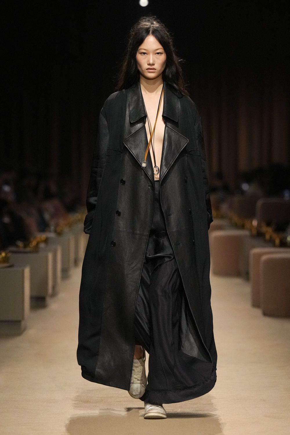 Oversized black coat, leather-faced front with broad peaked lapels. Two long tan cords hung with rough crystal pendants. Black satin wide-legs, pooling and flashing a soft crease. Off-white low-top sneakers, visibly creased across the toe. For crossing a hotel lobby barefoot in spirit, shoes still on.