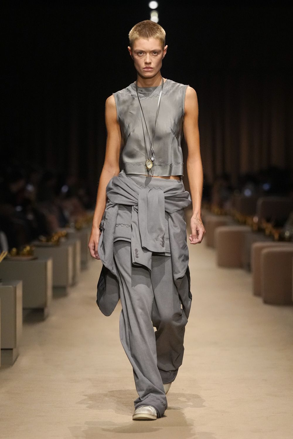 Pewter satin shell top with raw armholes and zigzag-joined seams. Long cord pendant necklace with a cloudy oval stone. Grey button-front shirt knotted hard around the hips. Slouchy grey trousers breaking over square white shoes. Square-toe white slip-ons. For crossing an empty hotel lobby before breakfast is served.