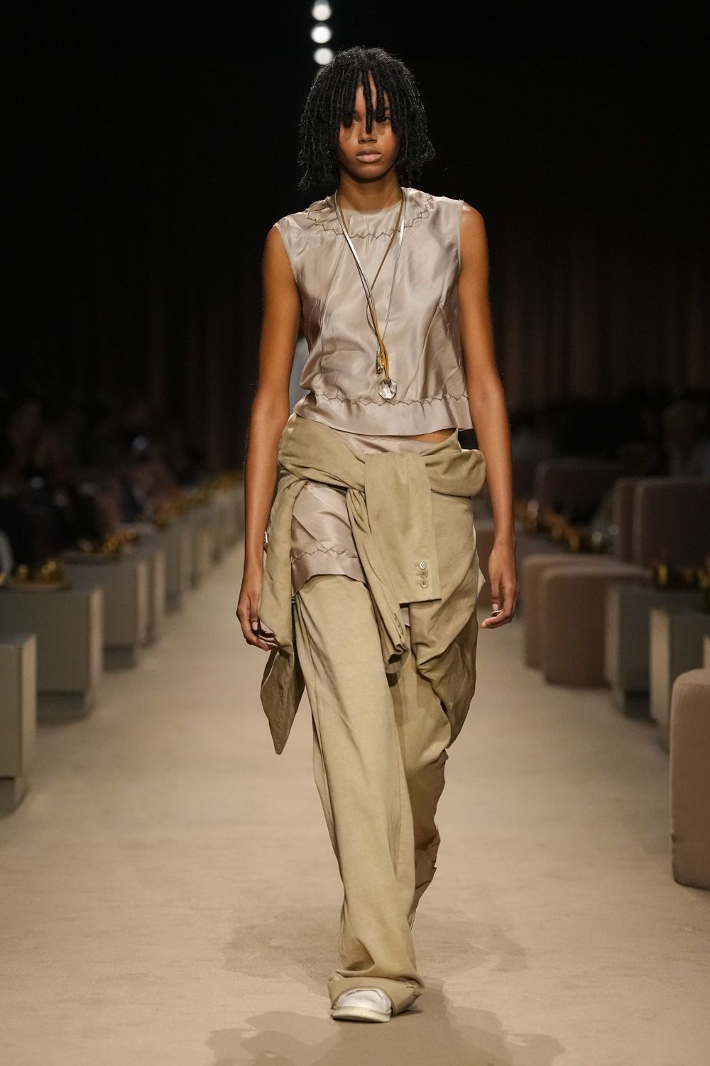 Pale dove satin shell, raw zigzag seams puckering the neckline and hem. Long cord necklace with a rough clear stone drop. Sand suede trousers, pooled wide over blunt square toes. Matching suede jacket knotted low at the hips, cuff buttons showing. For the desert motel checkout when the air is still cold.