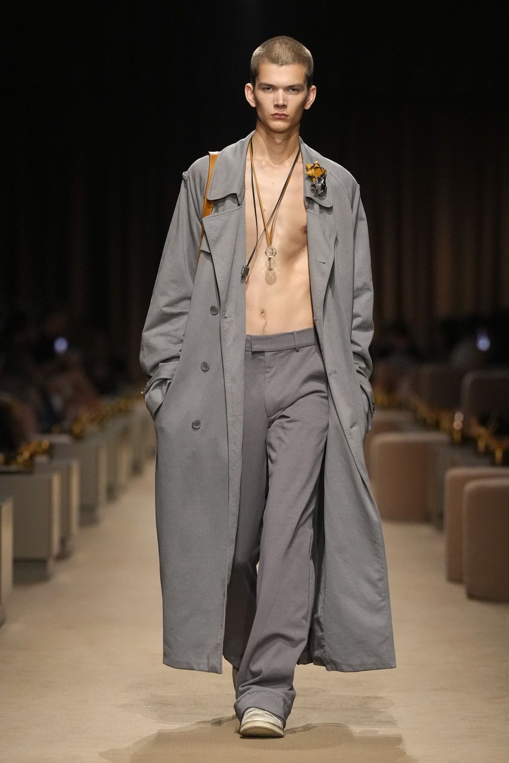 Slate cotton trench worn bare-chested, collar slouched and cuffs loosely belted. Tangling cords with a clear orb and a small metal fob. Crushed yellow rose pinned beside a crumpled silver wrap. Matching straight trousers breaking softly over white rubber-toe sneakers. For carrying flowers home after the apology already worked.