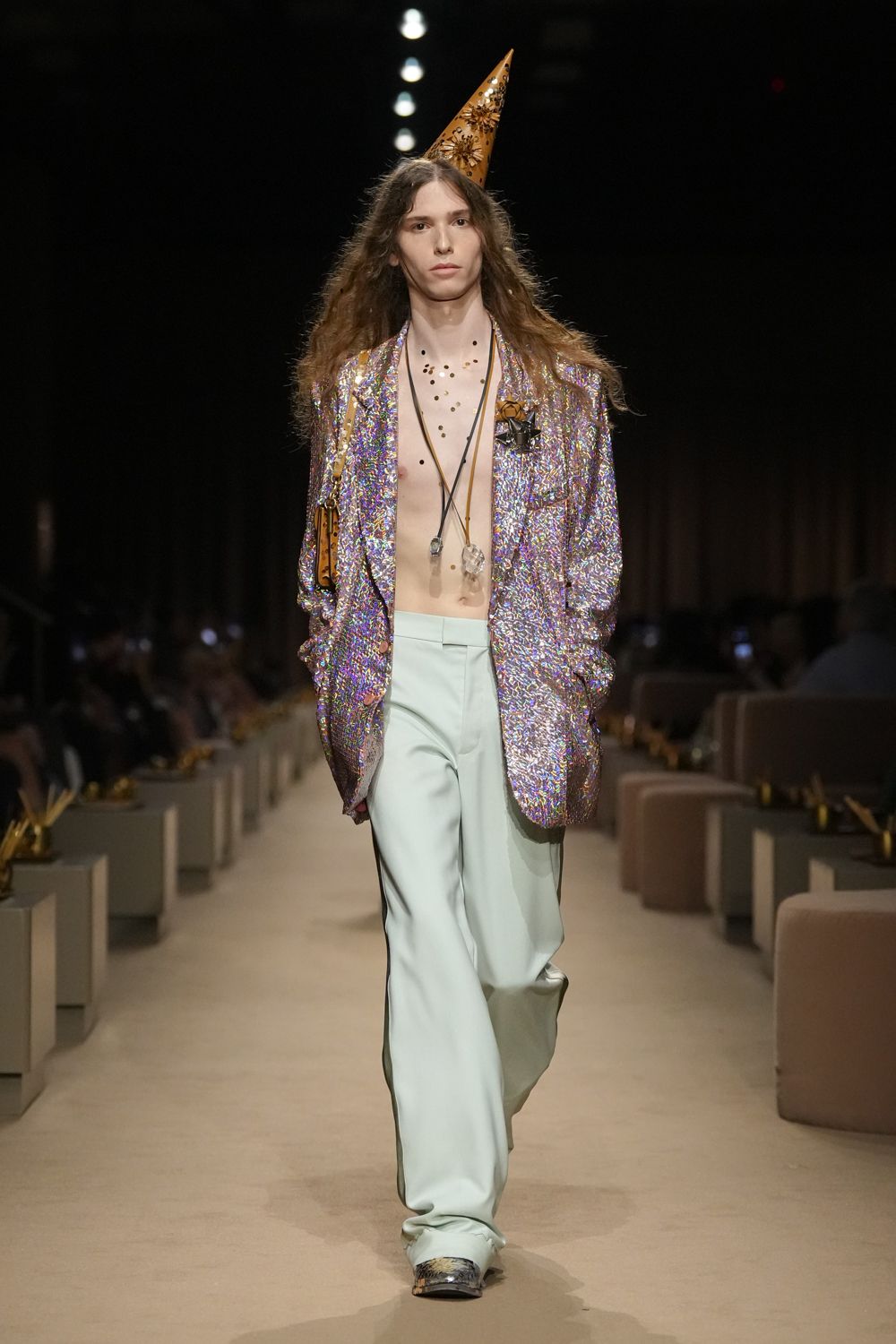 Tan leather cone hat punched with floral cutouts and studs. Oversized iridescent sequin blazer, a rosette pinned at one lapel. Long cords with raw crystal pendants over the bare chest. Pale mint trousers falling long over the shoe. Silver snakeskin square toes. After midnight at a birthday dinner where the cake arrived on fire.