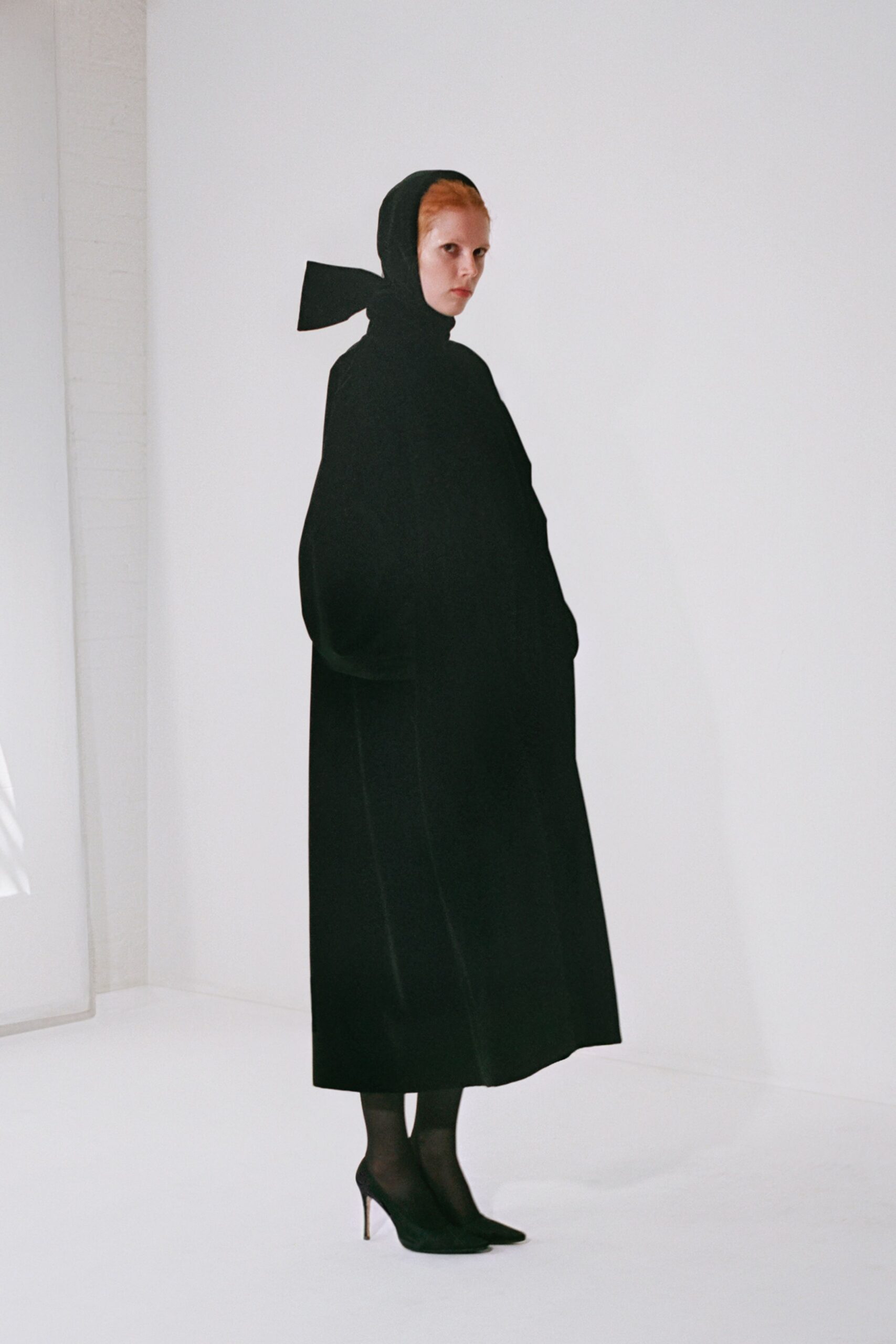 Black scarf tied low, one stiff tail jutting behind the head. Floor-length black coat in dense wool, cut with swallowed hands at the sides. Sheer black tights. Black pointed pumps with thin metal stiletto heels. Church steps in winter, after the bells and before anyone speaks.