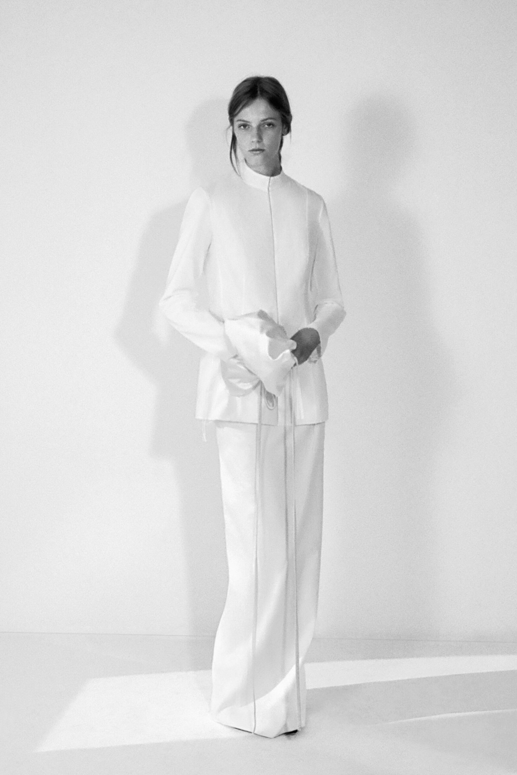 Ivory high-neck tunic with a concealed placket and long drawcord ties. Floor-length ivory trousers, pressed with a sharp crease through the toe. Square toes barely breaking the hem. Morning light in a registry office before anyone has found the rings.
