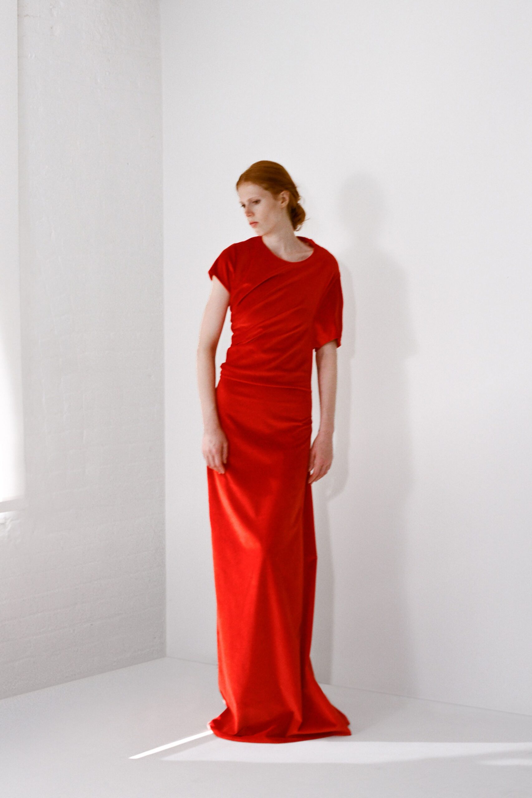 Scarlet column gown in fluid jersey, diagonally draped with one cap sleeve. Morning light in an empty loft before anyone has brought the chairs in.