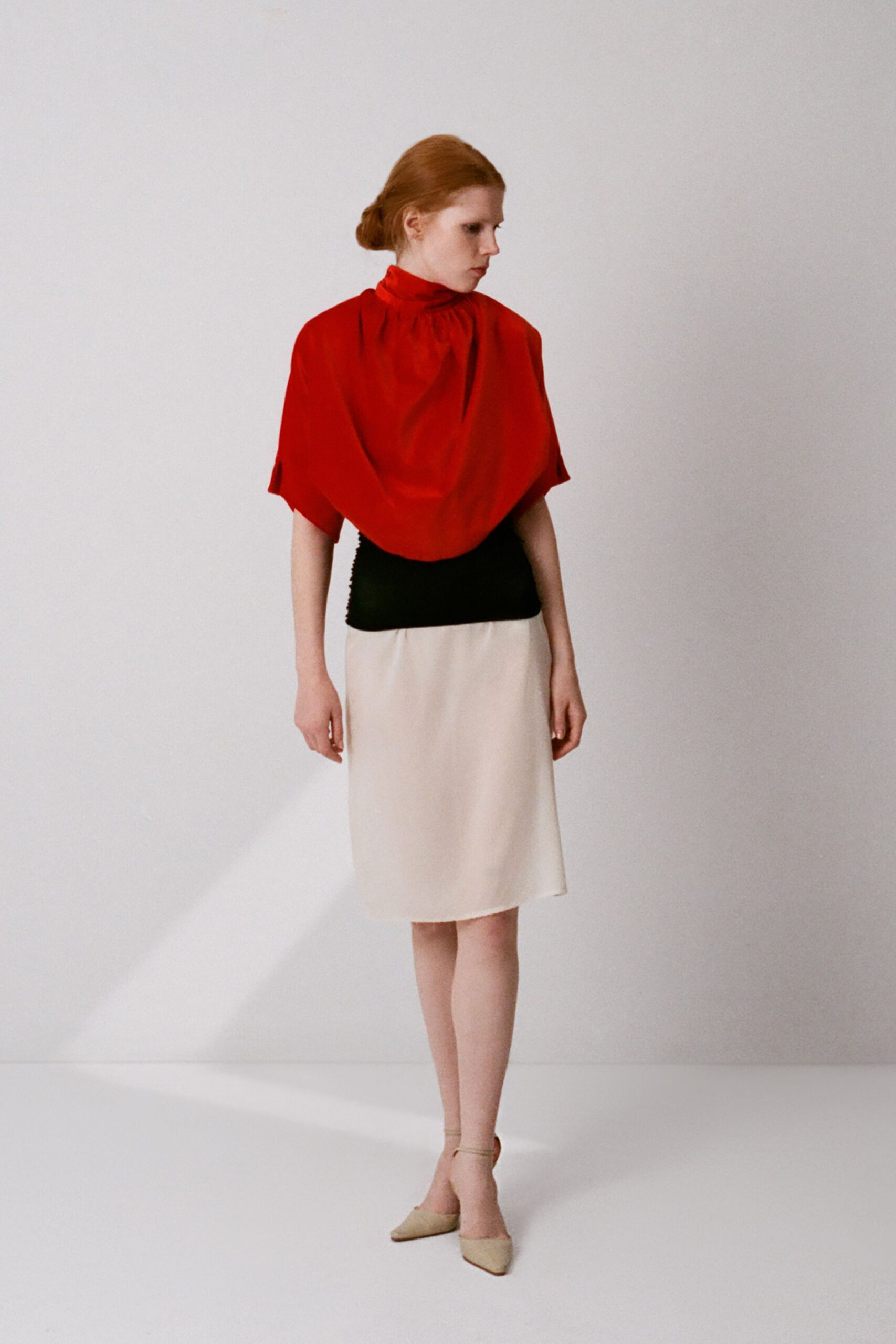 Tomato-red cape top with a gathered funnel neck and drooping front. Black ribbed knit panel hugging the midriff. Cream satin midi skirt cut straight, falling without a crease. Oat pointed pumps with thin ankle straps. Museum atrium at opening hour, still hearing the mop bucket roll away.