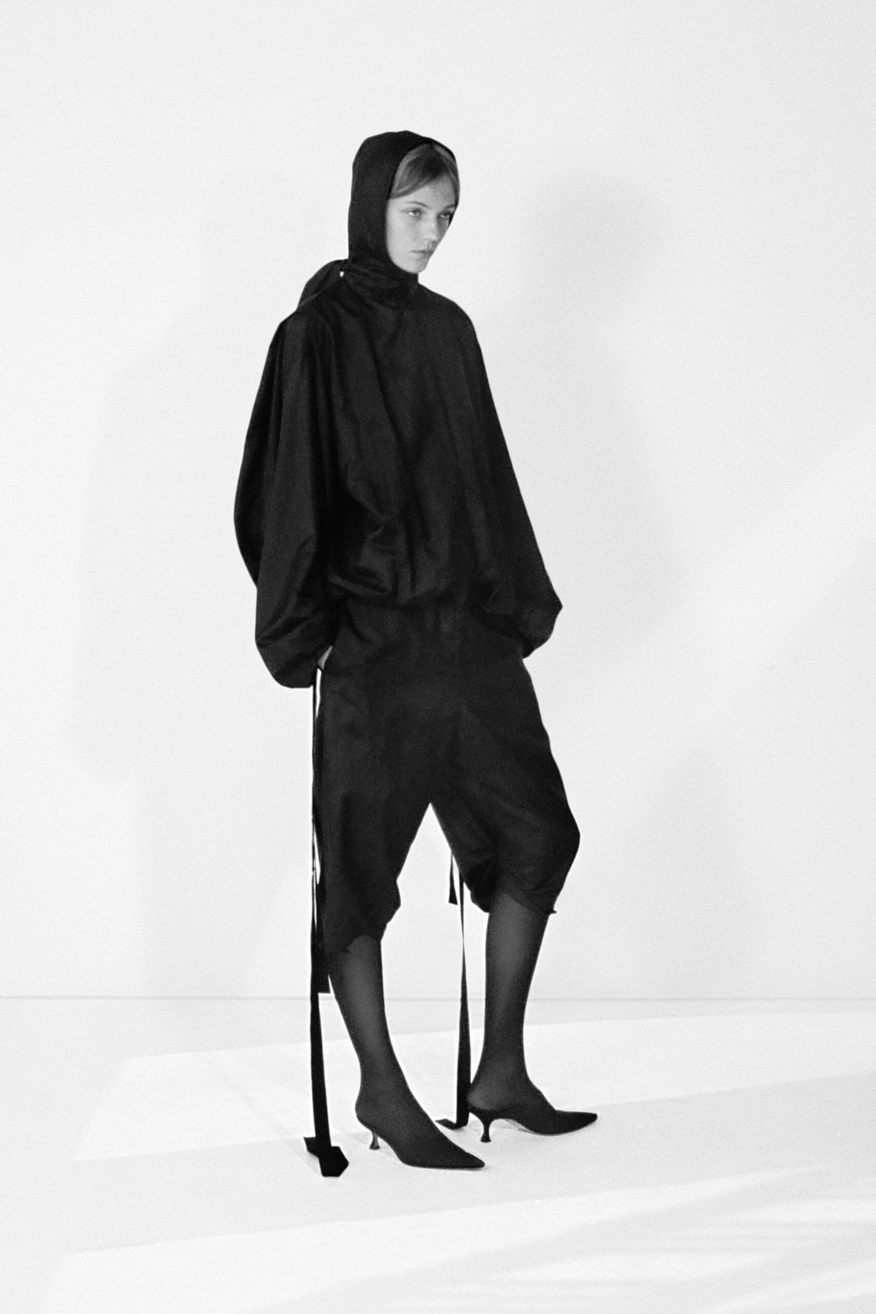 Black jersey hood wrapped close at the neck. Voluminous matte black shell blouson with ballooned sleeves. Loose black cropped trousers, raw hems caught just below the calf. Long black ties hanging past the ankle. Knife-point sock boots on pin-thin kitten heels. For crossing a silent stage after everyone else has left.