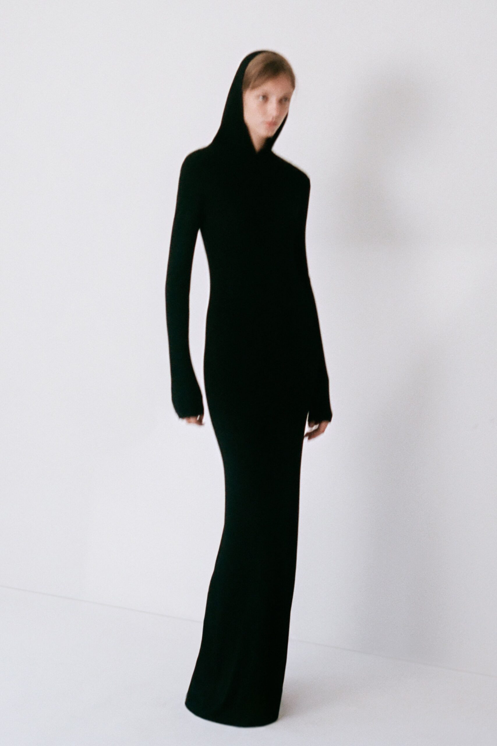 Black column knit, hood rising straight from a close funnel neckline. Extra-long narrow sleeves, fingertips just slipping past the cuffs. First train out after a sleepless winter blackout.