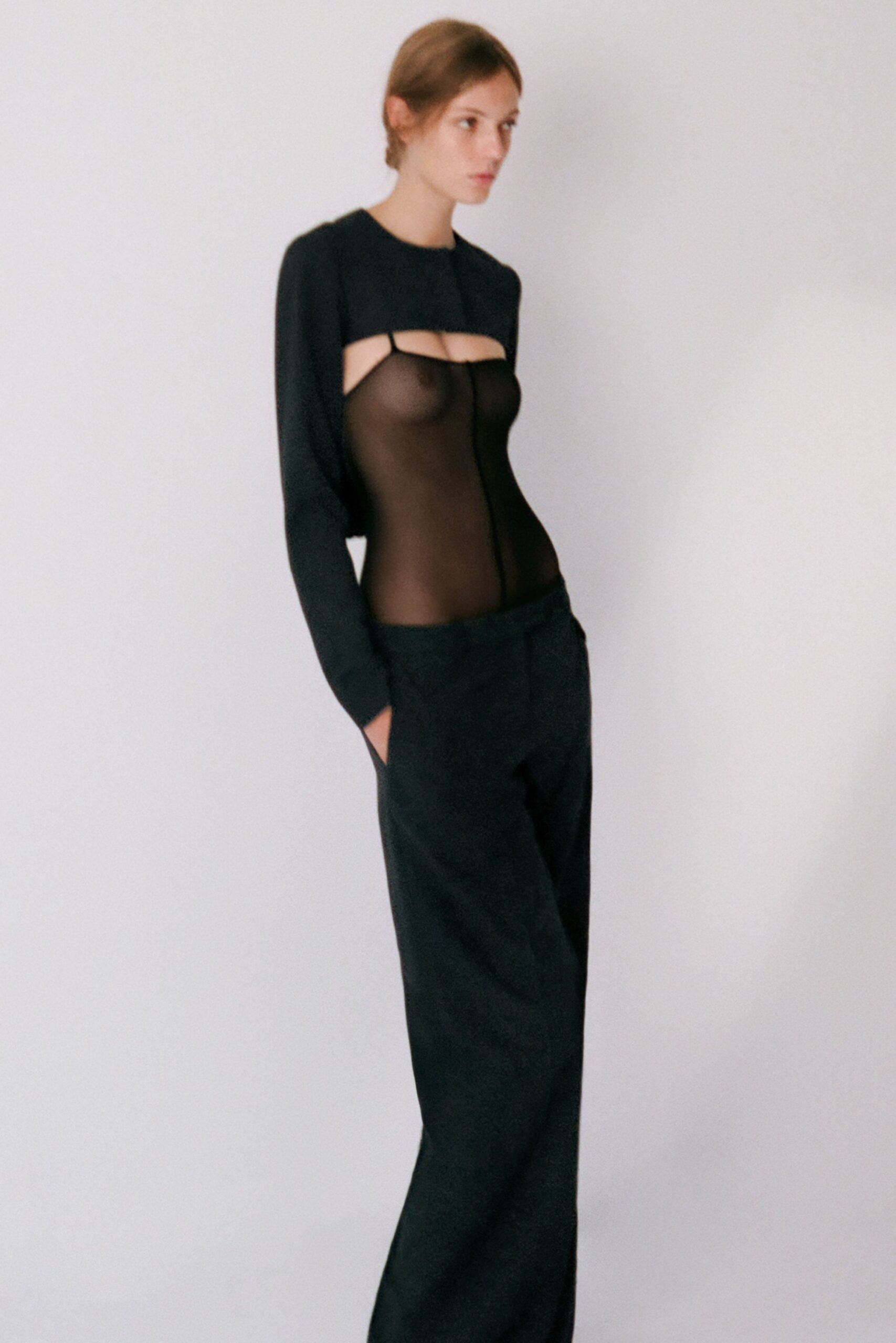 Black long-sleeve crop top, cut high under the bust with slim straps. Sheer black mesh bodysuit, center seam running straight down the torso. Low-slung black trousers with loose straight legs and hands-deep pockets. Whatever you wear home when dawn finds you still awake.