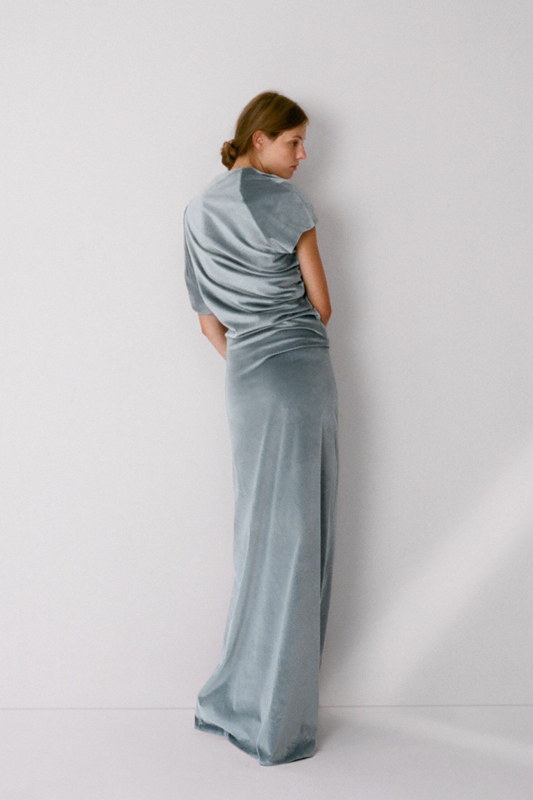Pale blue velvet top, liquid drape gathering into twists at the side. Matching floor-length velvet skirt, pooling softly over the feet. At the museum after closing, still catching the last cold light.