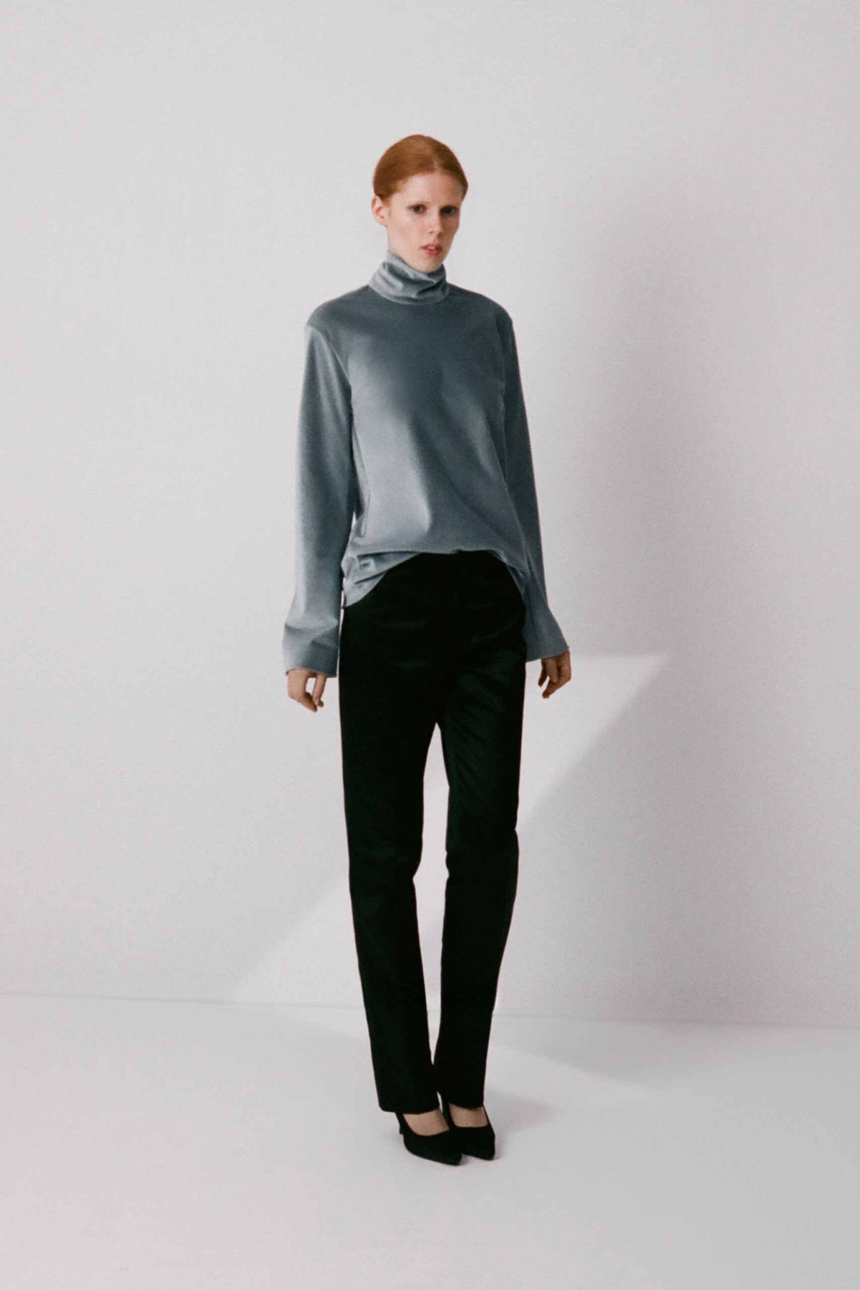 Pale blue satin jersey turtleneck, hem twisting slightly off center. Black slim trousers with a faint pressed crease. Black suede pumps. Early appointment on the quiet floor of a private clinic.