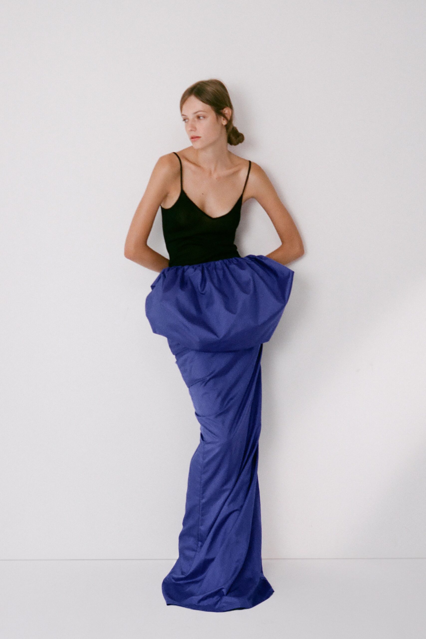 Black knit camisole bodice with narrow straps and a scooped neckline. Cobalt taffeta column skirt, gathered into a ballooned hip shelf and trailing hem. Opening the apartment door with the party still audible downstairs.