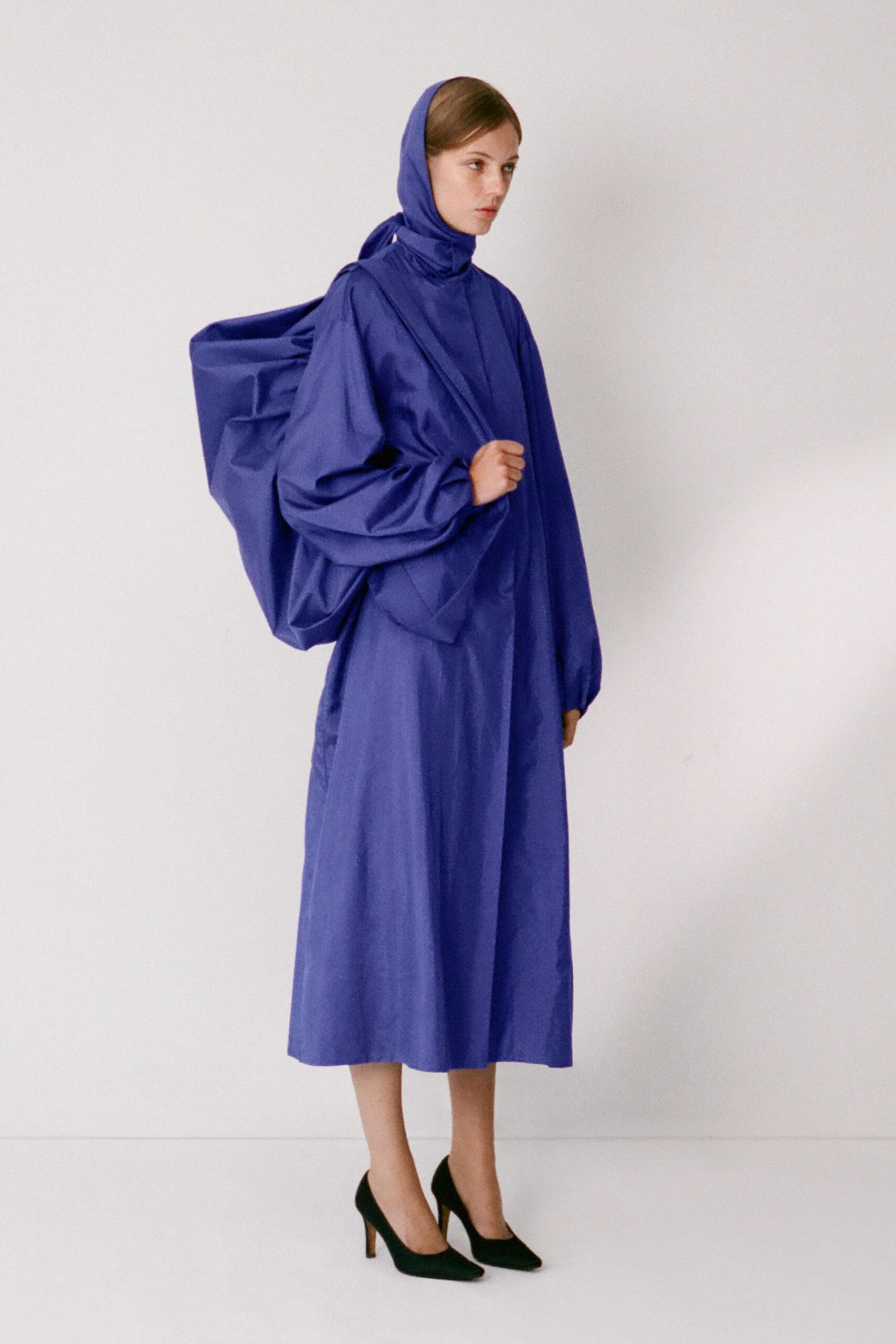 Cobalt cotton headscarf knotted low, wrapping close at the throat. Matching poplin midi dress with dropped shoulders and gathered stand collar. Oversized soft tote collapsing into square folds against the back. Black pointed pumps with slim heels. Crossing a windy ferry deck with your whole weekend in one bag.