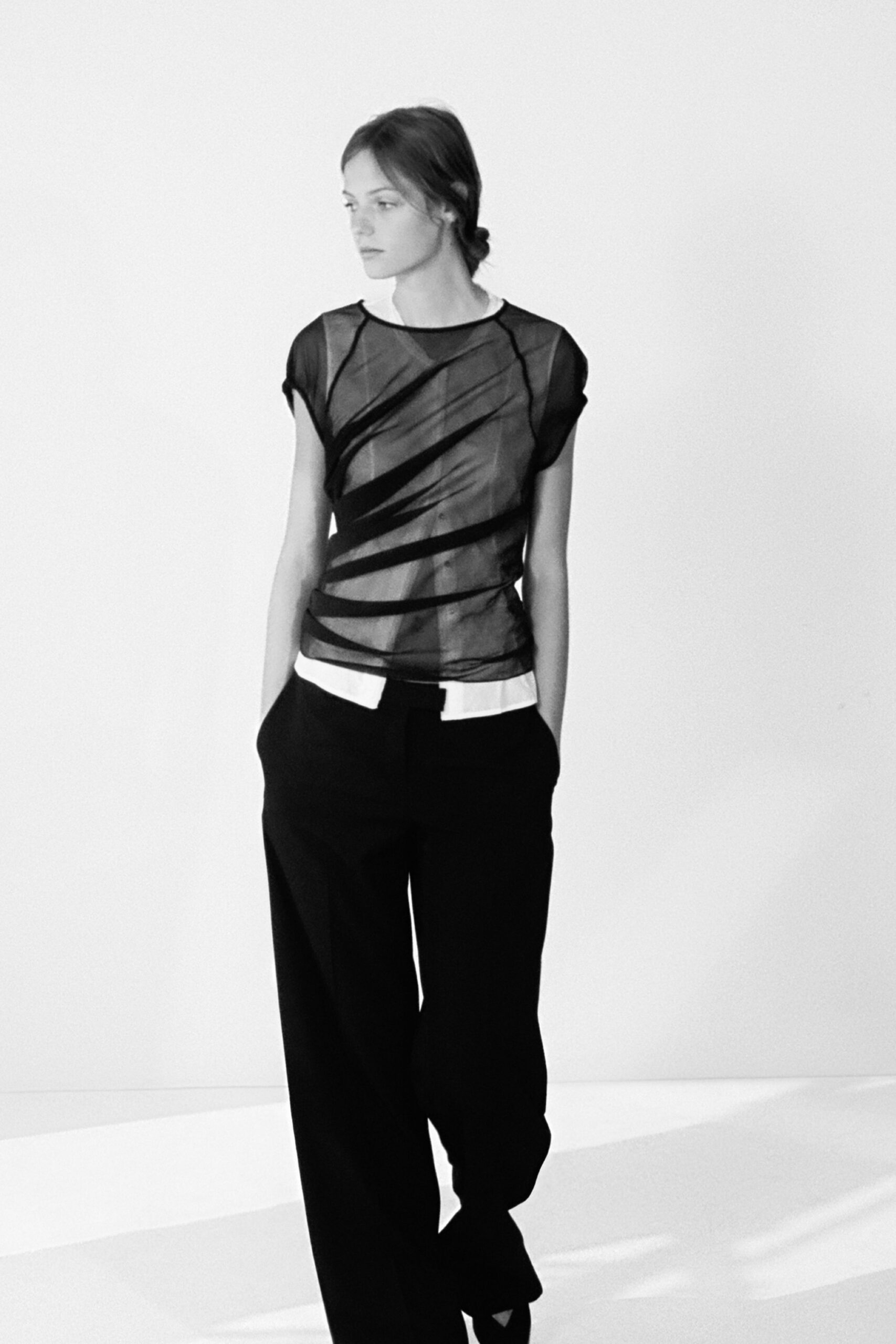 Black mesh cap-sleeve top, diagonal opaque bands twisting across the torso. White jersey peeking out in two tabs below the hem. Black straight-leg trousers with hands-buried side pockets. Walking home after a matinee, ticket stub still in your coat pocket.