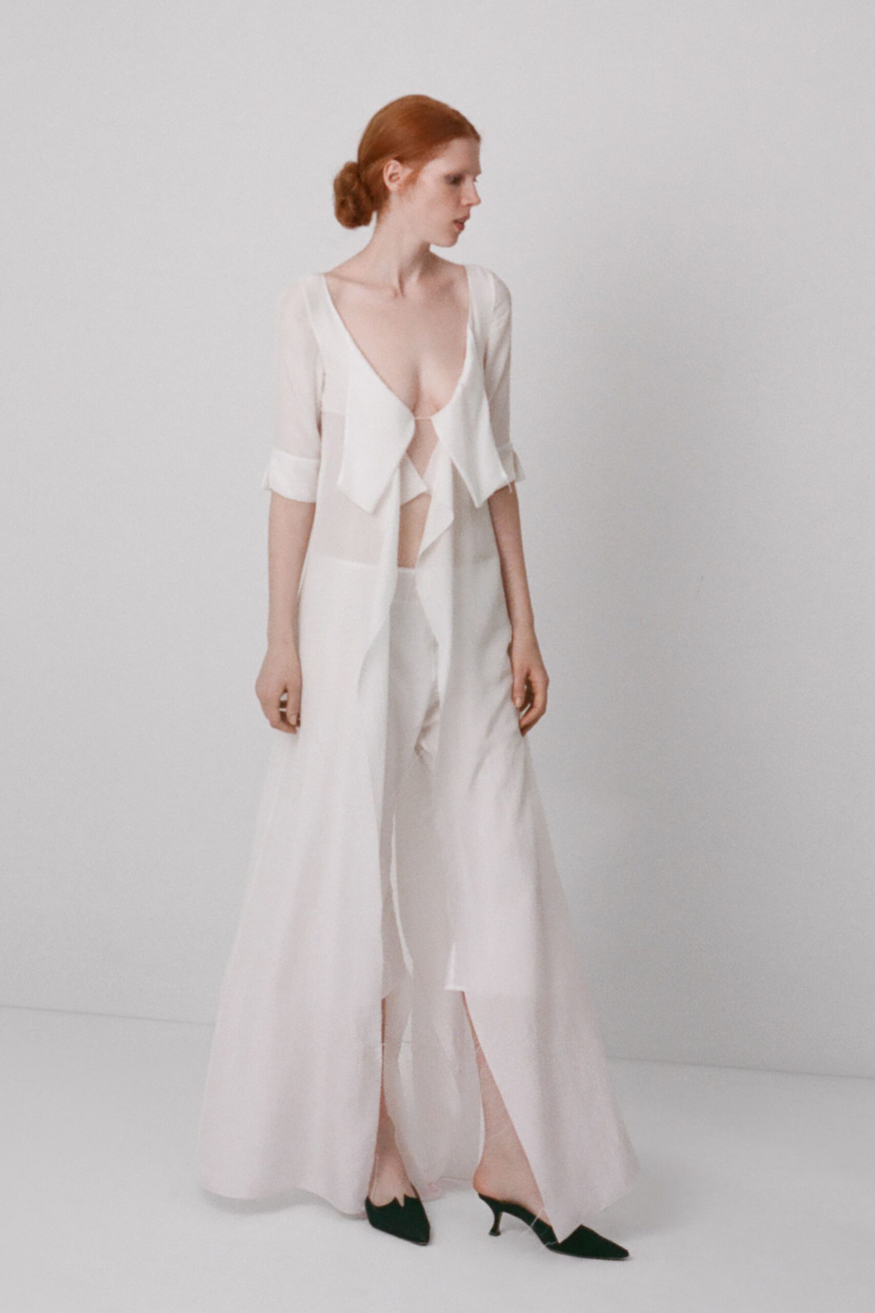 Sheer ivory duster falling to the floor, cuffs turned in a wide band. Plunging ivory top with scarf-like panels hanging loose at the center. Wide ivory trousers, soft and fluid under the transparent layer. Black pointed pumps with a sharply notched vamp. Crossing a hotel lobby at dawn before anyone has reset the flowers.
