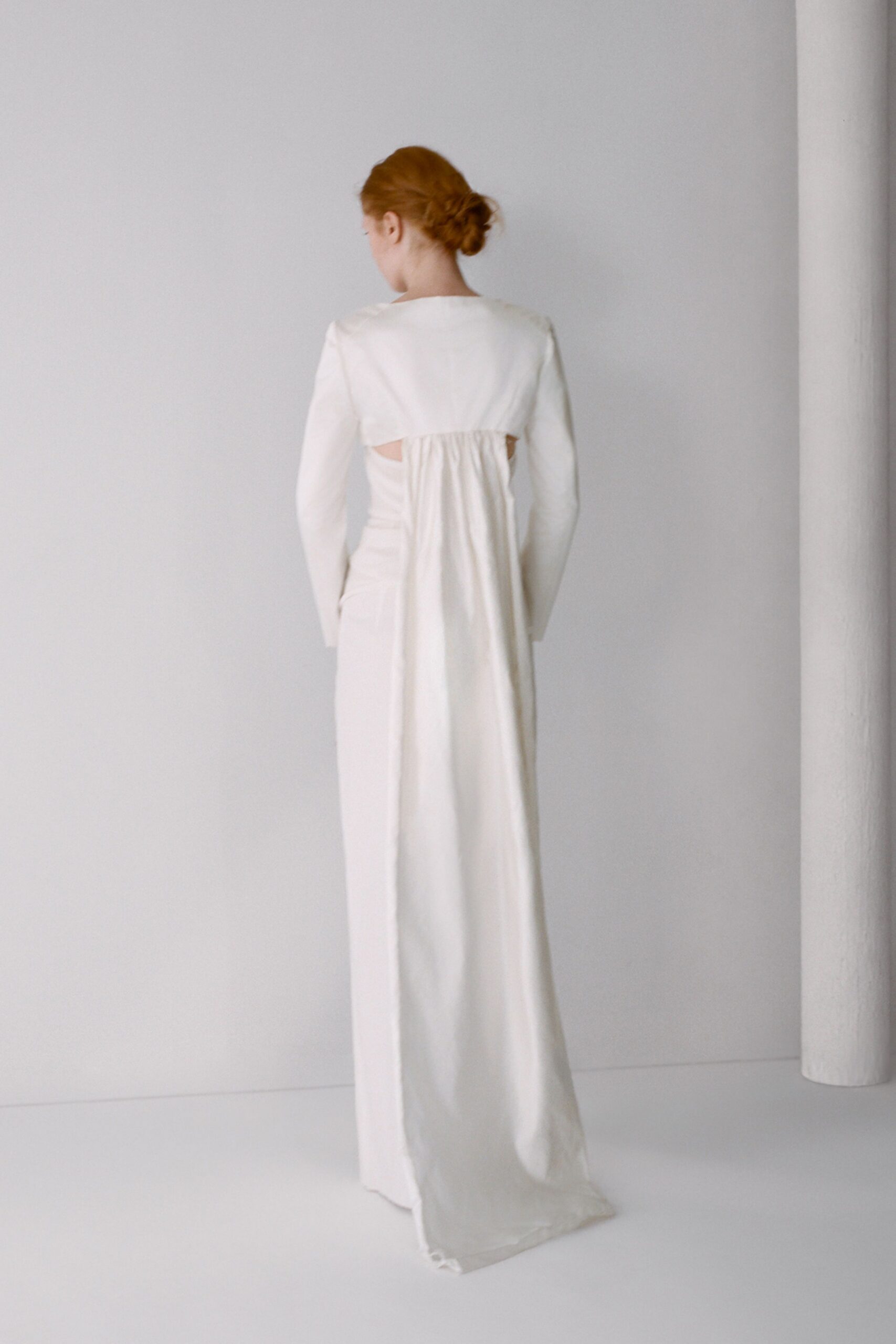 Ivory floor-length gown with a gathered back panel falling from a yoke. Long fitted sleeves and side cutouts exposing the ribs. At the registry office after everyone else has gone home.