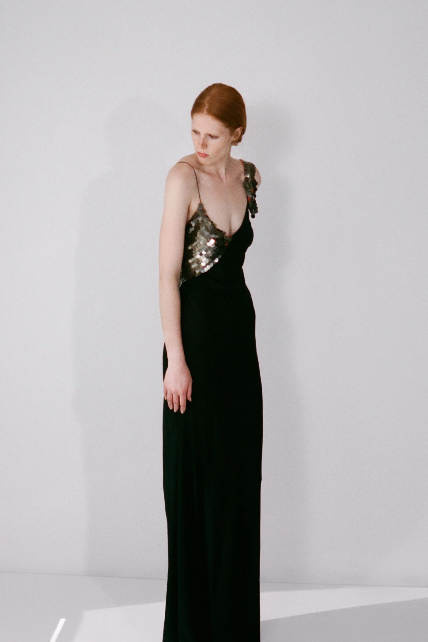 Black floor-length slip gown with a plunging V and gunmetal paillettes climbing one strap and bodice. Morning after a wedding, standing barefoot in the rented house.