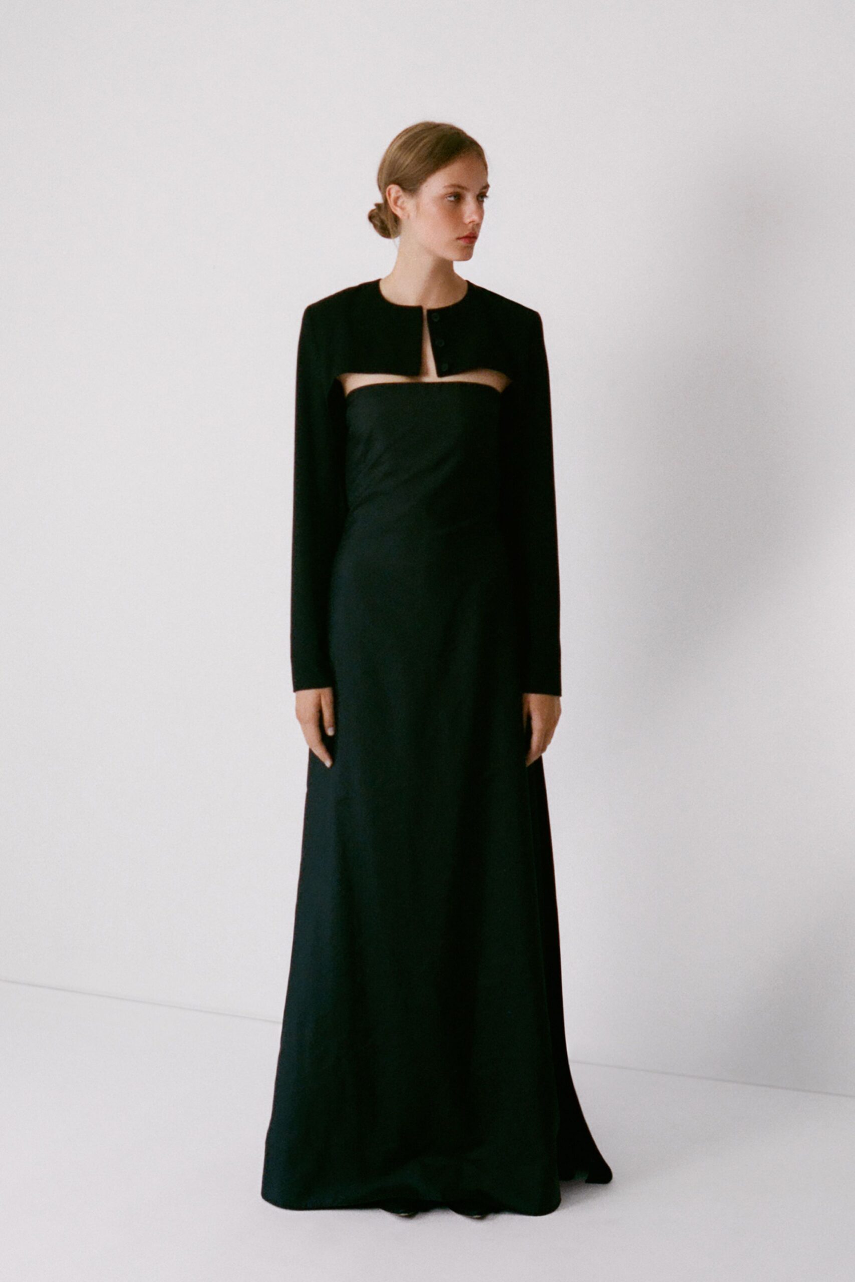 Black cropped bolero with a collarless edge and two small buttons. Strapless black column gown, cutout band skimming the collarbones above a fluid skirt. For slipping into a civil ceremony with rain still drying on the steps.