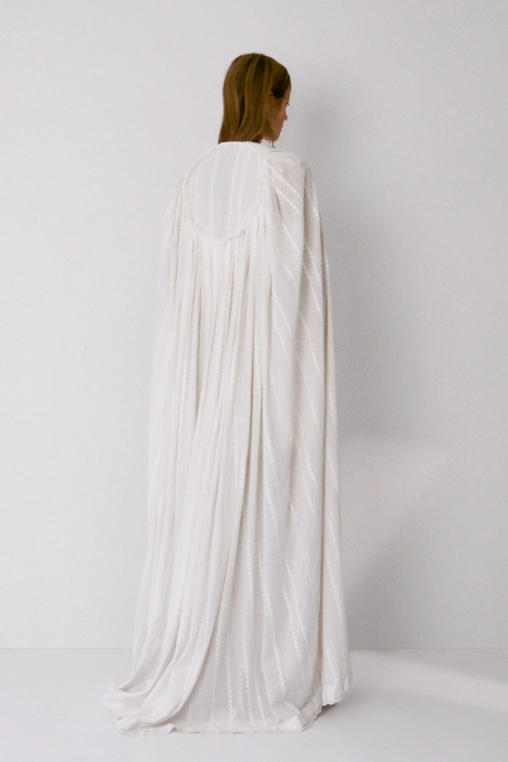 Ivory satin cape gown with fine vertical pleats falling from a rounded yoke. Floor-pooling hem, one back corner slightly kicked under. For crossing a marble hall after the ceremony has emptied out.