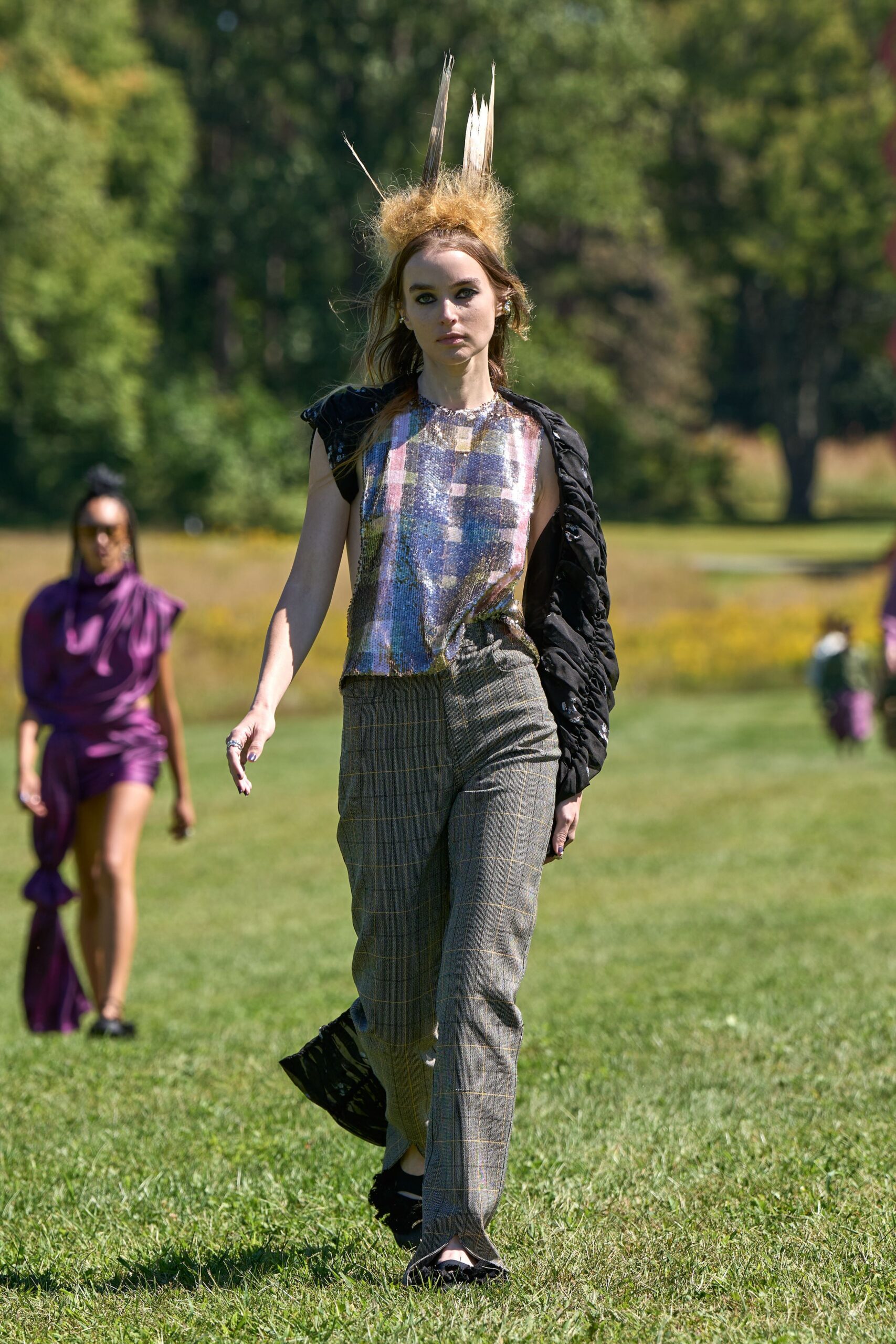 Tufted straw headpiece pierced with three upright feathers. Sleeveless sequined shell in blurred mauve and blue checks. Black ruched jacket shrugged off one shoulder. Grey glen plaid trousers slit open over the toes. Black pointed flats. Crossing a sculpture park after the lawn has just been cut.