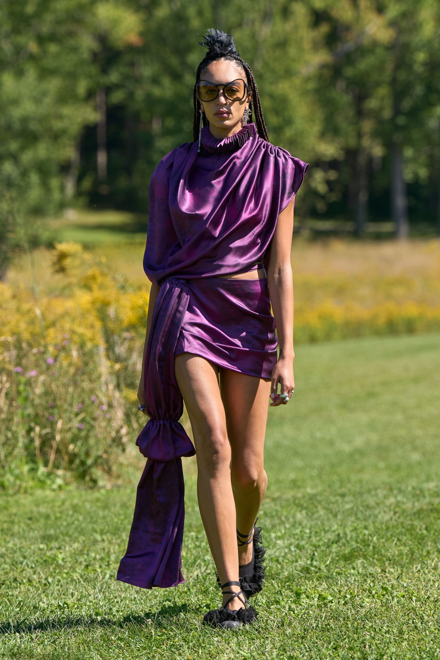Oversized amber-tinted lenses in a rounded black frame. Aubergine satin mini dress, ruched collar and one gathered balloon sleeve. Long sash train cinched near the knee, swinging from the waist. Black wraparound flats with shaggy ruffles at the toe. Crossing a meadow toward lunch on the grounds after rain.