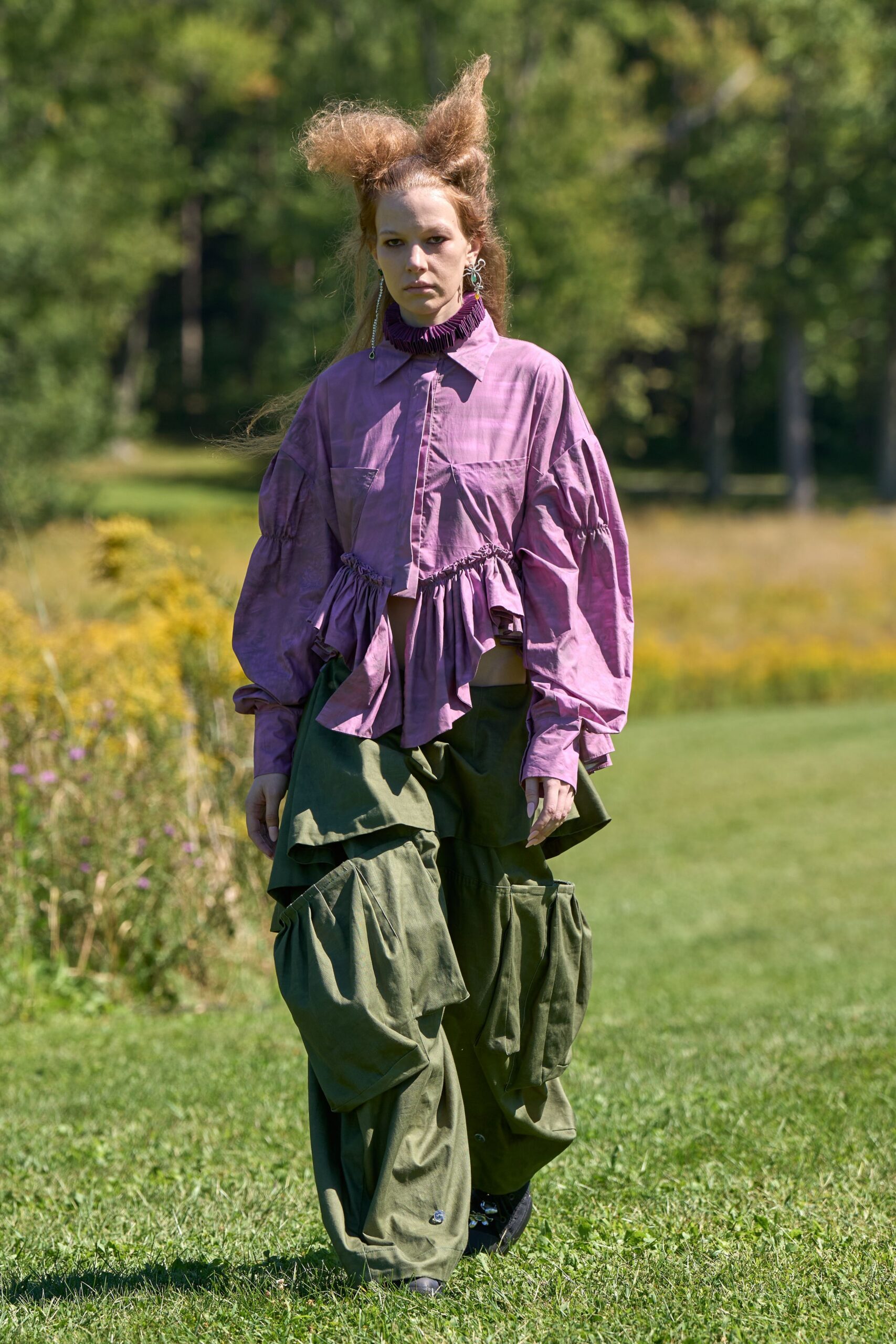 Plum pleated ruff packed tight around the throat. Orchid cotton blouse with dropped sleeves and a jagged gathered peplum. Olive parachute trousers ballooning into oversized flap pockets at the thigh. Black shoes half-hidden under the dragging hem. Crossing a mown field after rain, earrings still catching the sun.