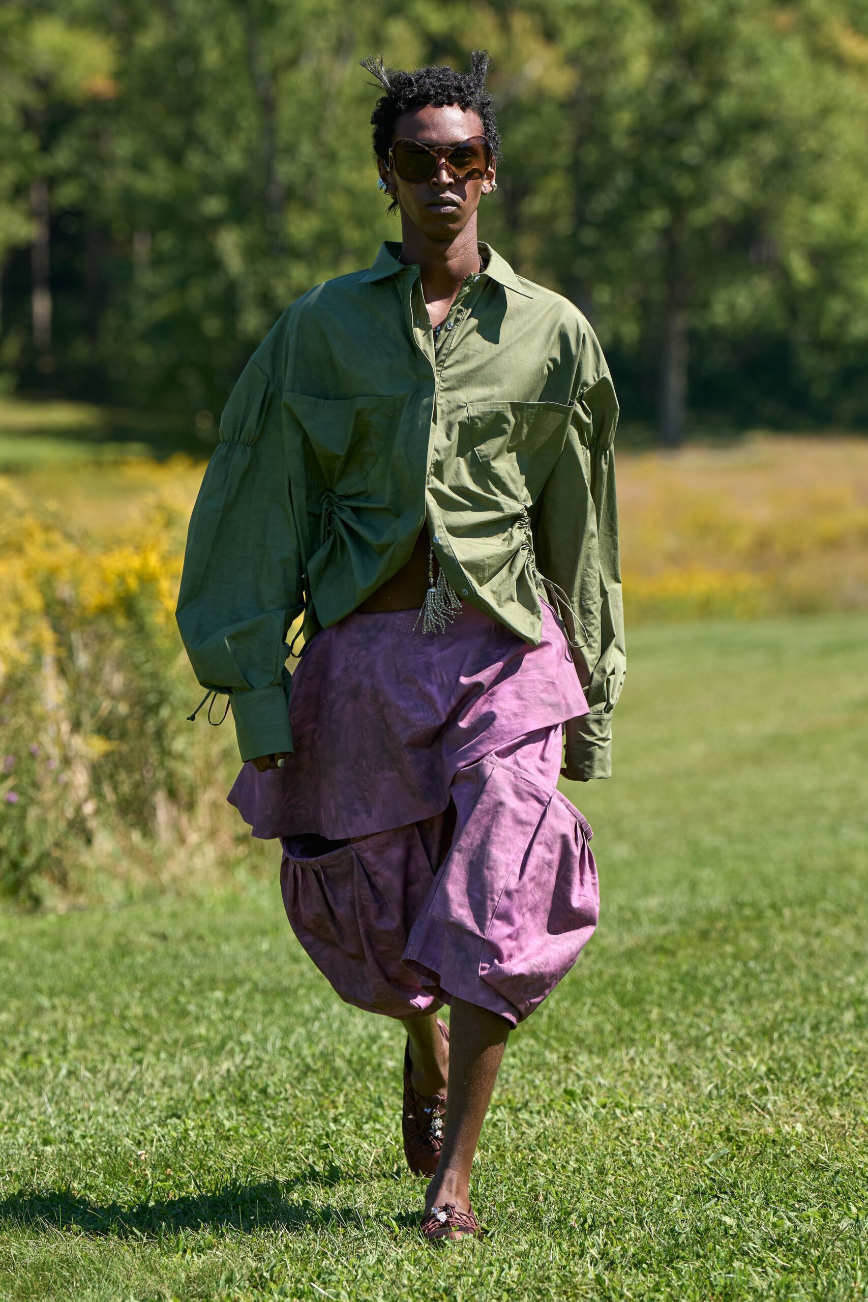 Oversized brown shield lenses with a softly squared rim. Olive poplin shirt, side seams ruched on dangling drawcords. Mauve taffeta skirt with a wrapped overskirt and ballooned underpanels. Brown lace-up flats, jeweled at the vamp. Walking across a meadow after rain, hem catching seedheads.