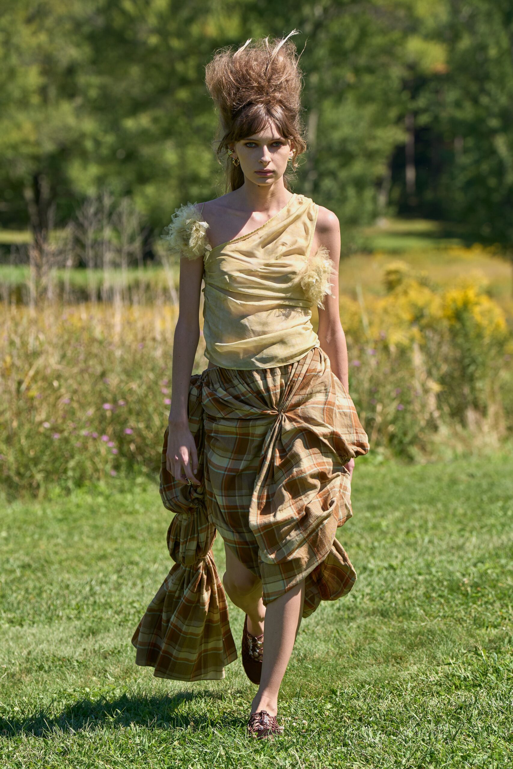 Butter-yellow one-shoulder top in crinkled voile, ruched across the chest. Pale tulle pompons clustered at one strap and underarm. Caramel plaid wrap skirt, twisted and knotted into heavy side swags. Burgundy woven leather flats with thin ties crossing the instep. Walking across a lawn after lunch, grass still damp at the hem.