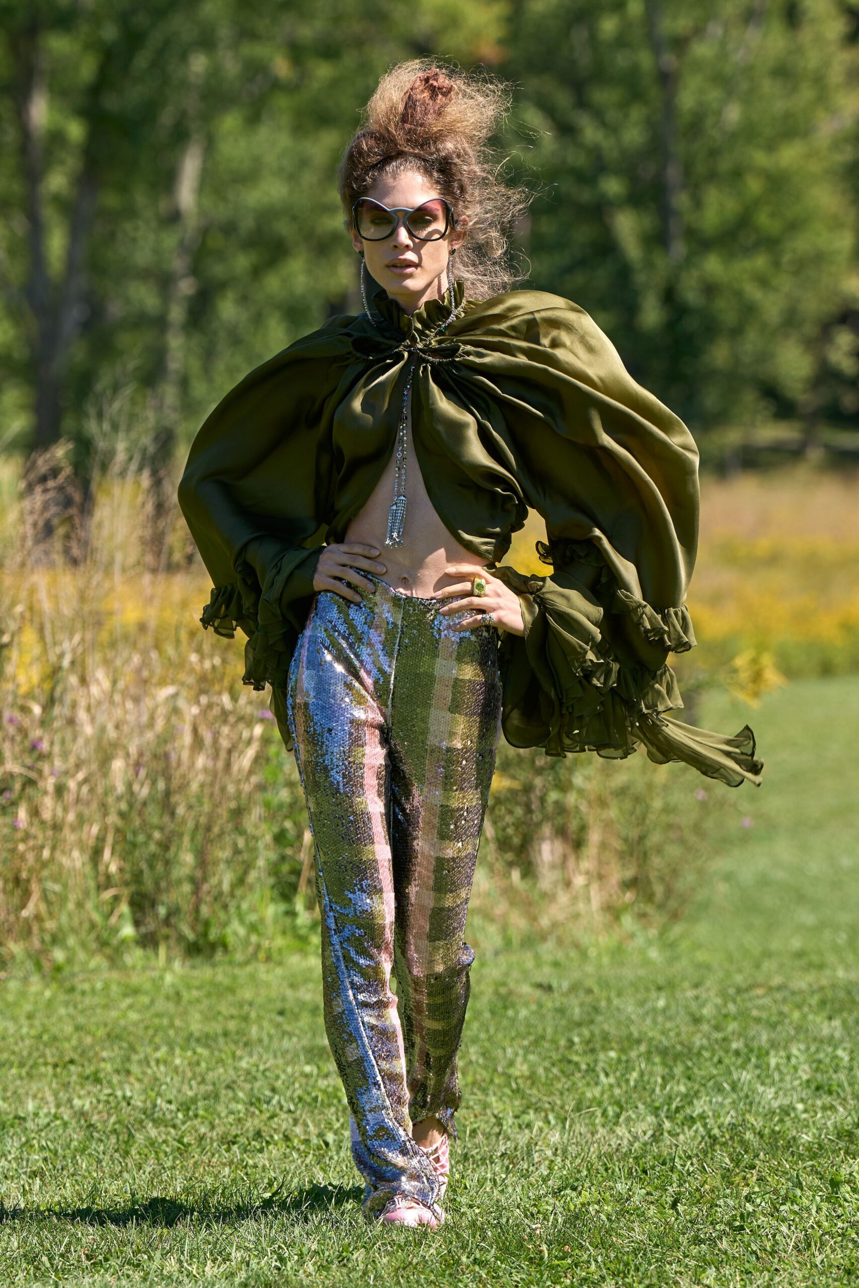 Oversized black frames with a soft butterfly curve. Olive satin cape blouse, neck shirred tight with a rhinestone tassel. High-waisted silver sequin trousers catching pink and meadow green. Pale pink lace-up sandals. Walking across a country lawn after the champagne has gone warm.